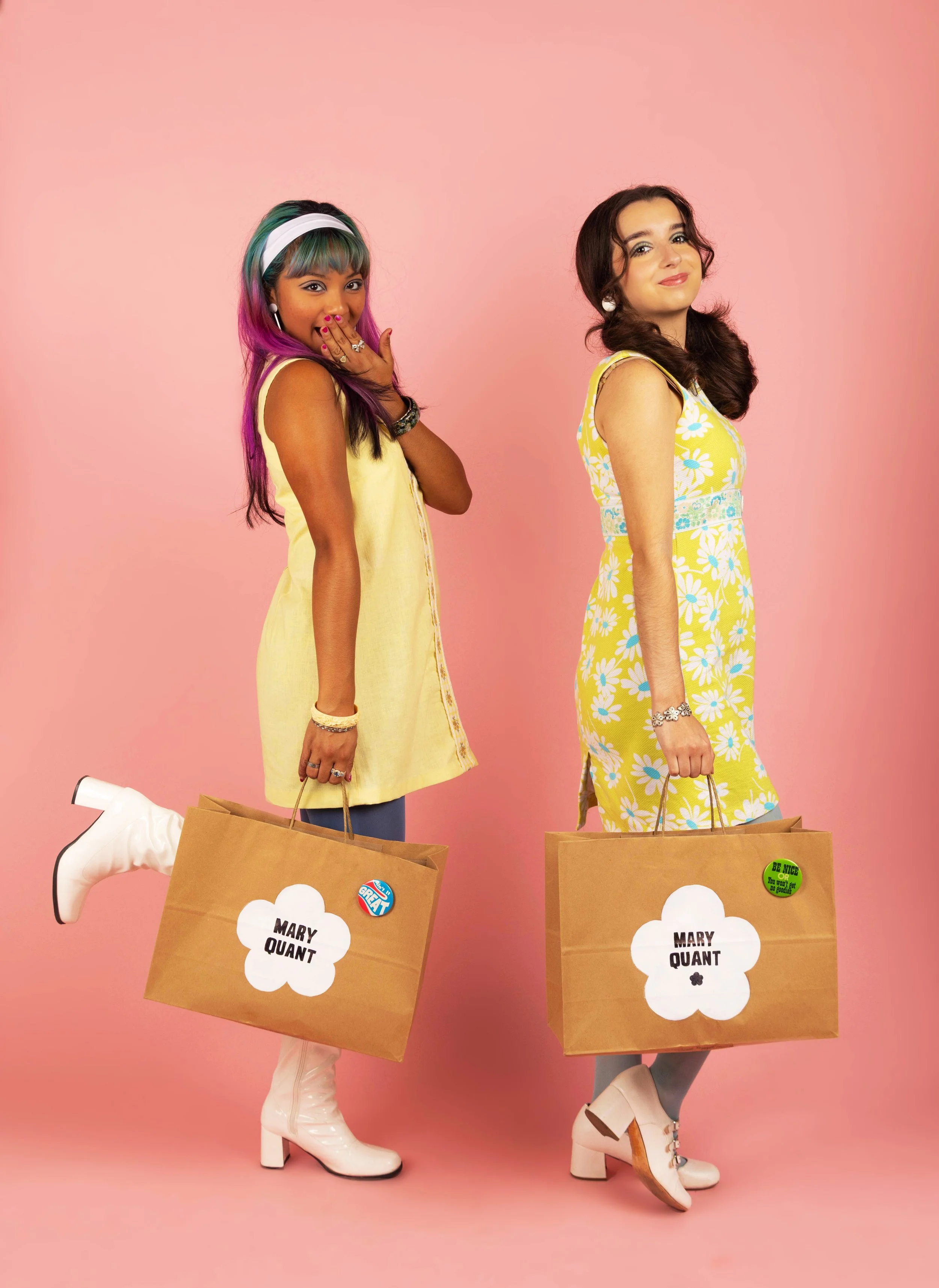 Two young women standing against a pink background, holding large shopping bags with the name 'Mary Quant' on them. They are dressed in colorful, vintage-inspired clothing and wearing platform shoes.