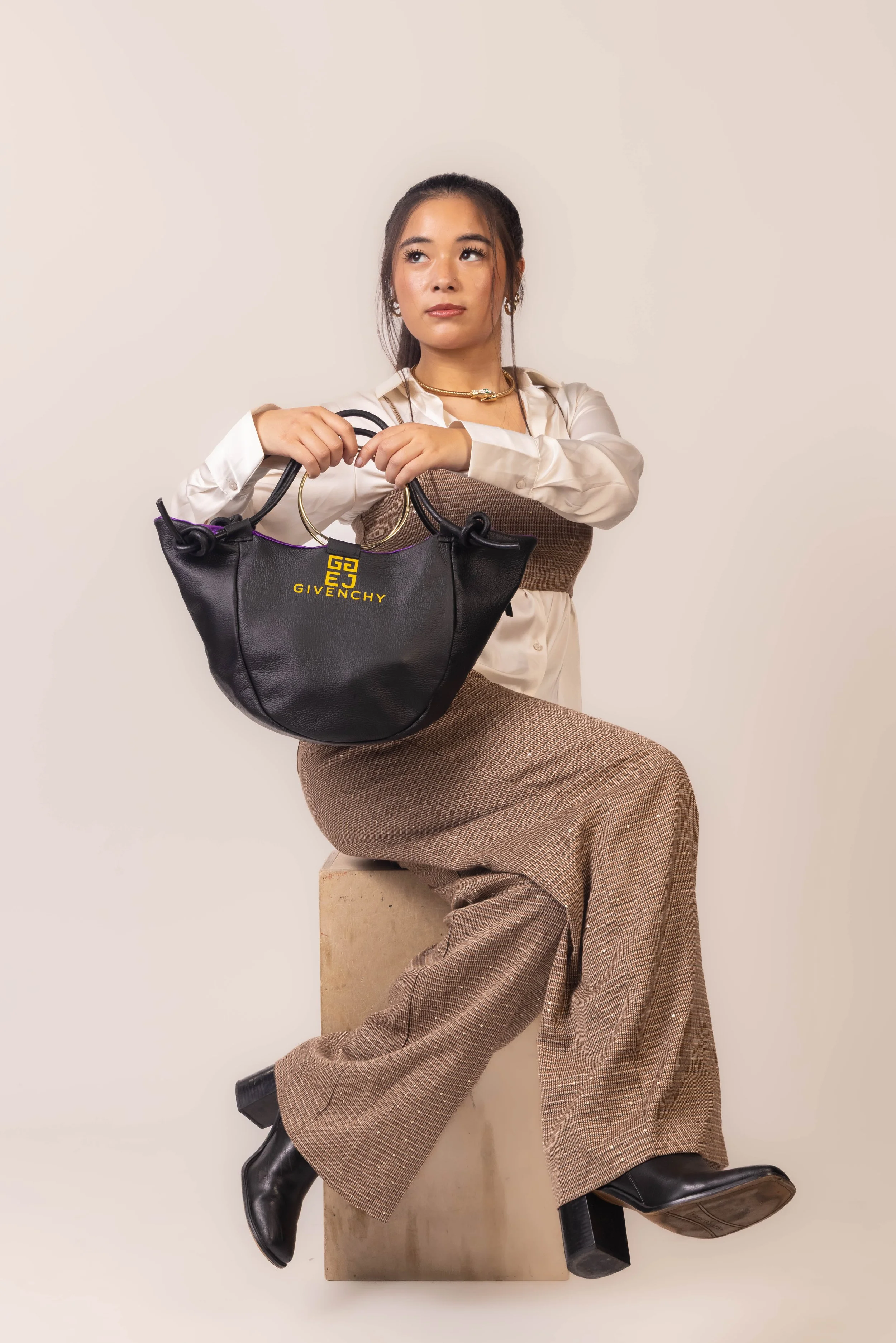 A woman with dark hair tied back, wearing a cream-colored blouse and brown houndstooth trousers, seated on a beige block against a plain background. She holds a black Givenchy handbag with gold branding and gold rings, and wears black boots and gold 
