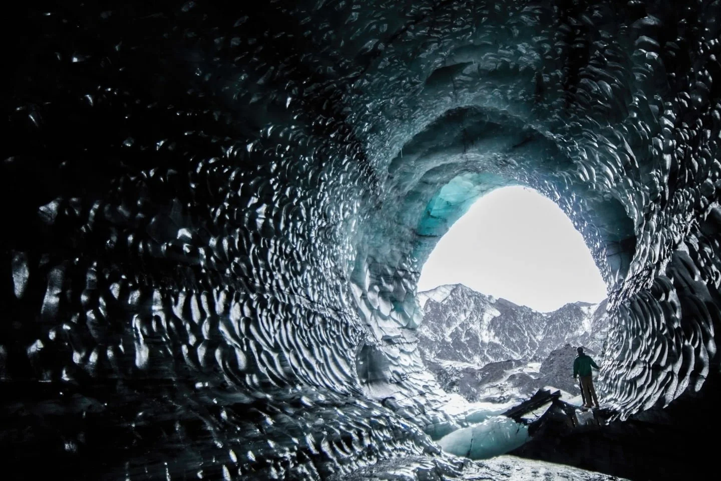 A person standing inside a large ice cave with jagged, textured ice walls and snow-covered mountains visible through the cave opening.