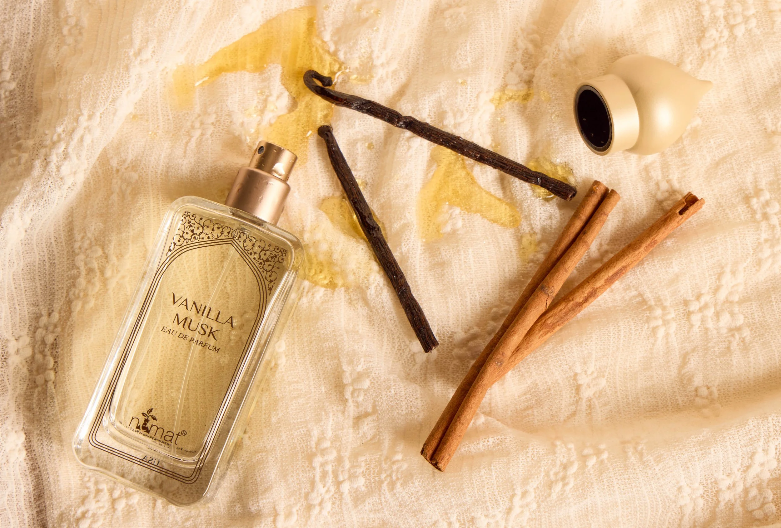 A flat lay of vanilla musk perfume bottle, two cinnamon sticks, two vanilla bean pods, and a white container of vanilla extract on a lace fabric surface.