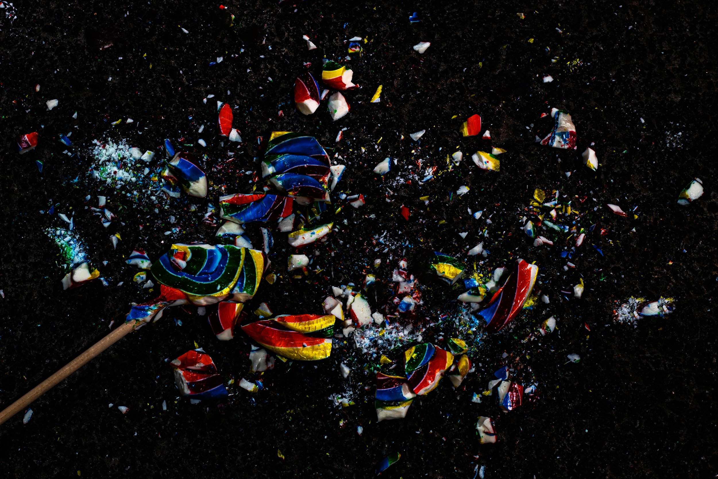 Broken colorful rainbow swirl lollipop pieces scattered on a dark black surface with a wooden stick attached.