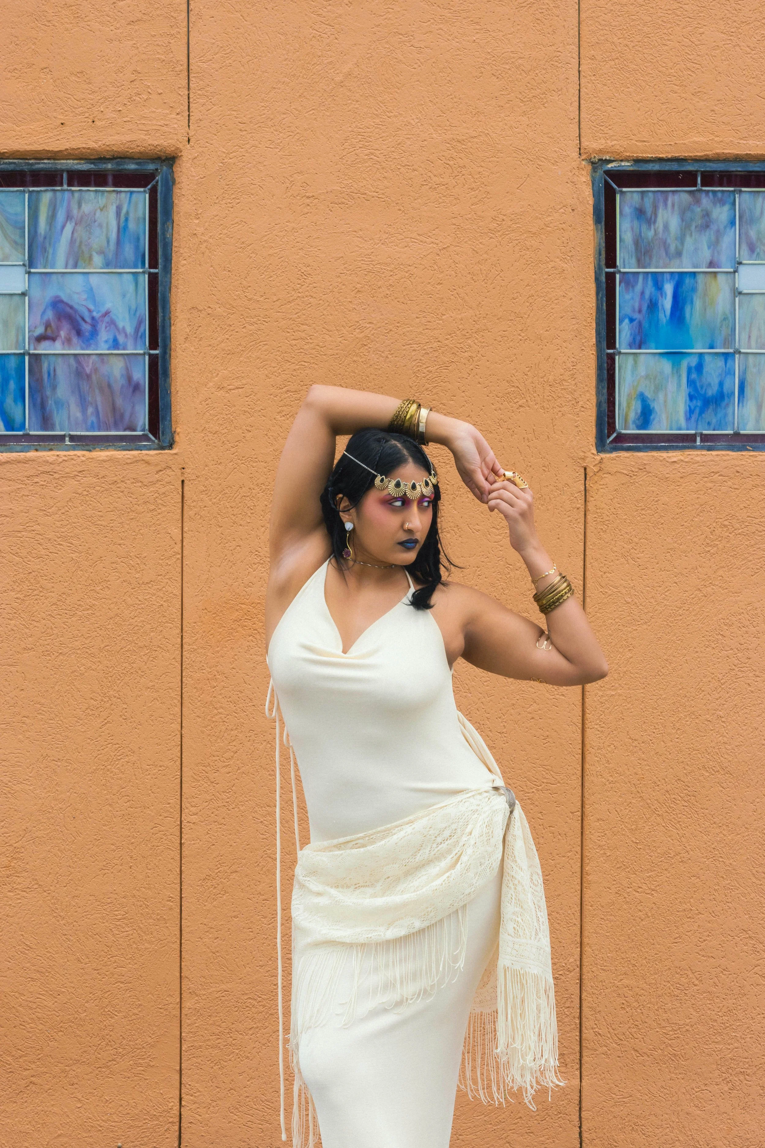A woman with dark hair and colorful makeup, wearing gold jewelry and a flowing off-white dress, standing against a textured orange wall with two stained glass windows.