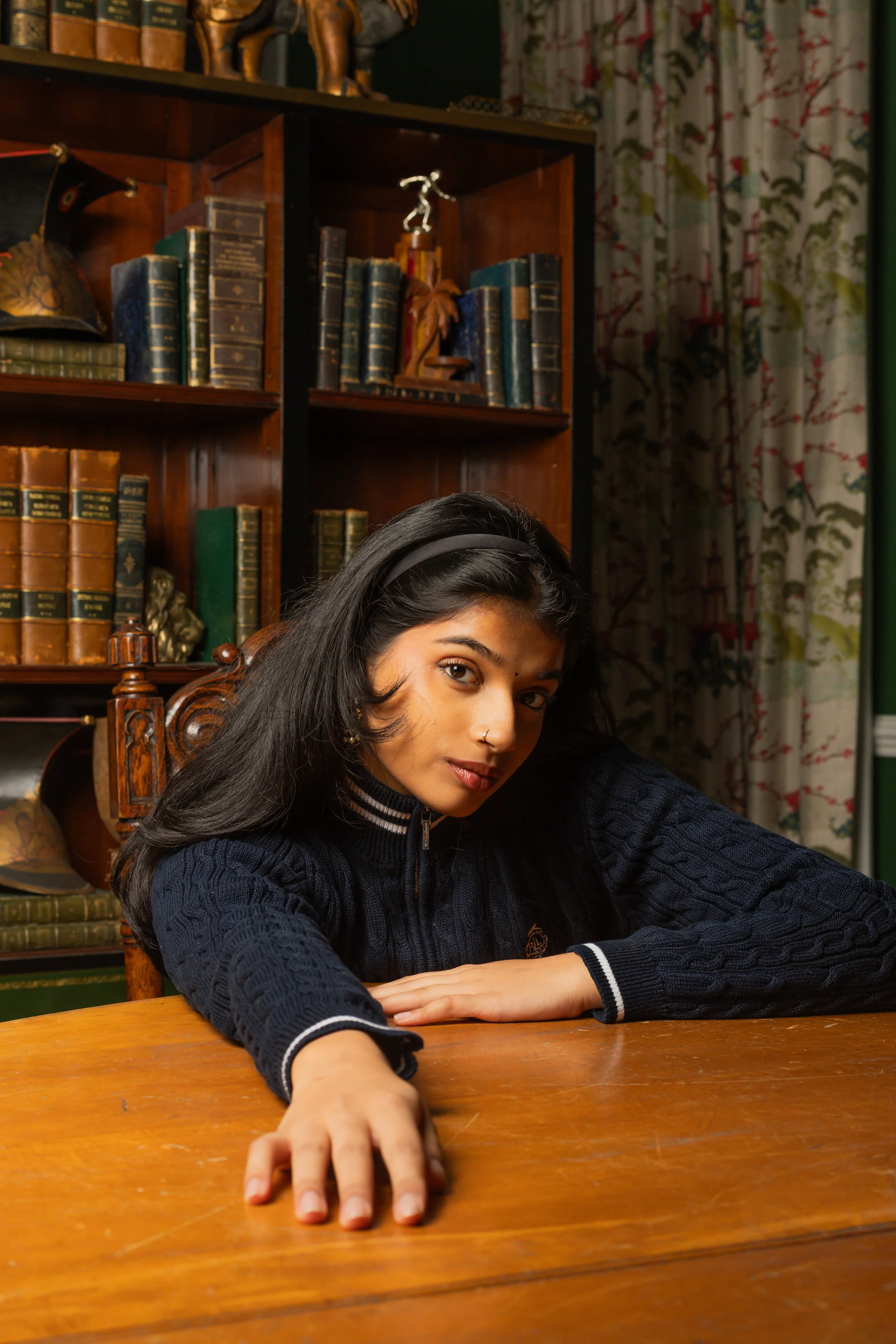 Portrait of a young woman with black hair and nose piercings sitting at a wooden table, reaching out with her arm extended, in a room with a bookshelf filled with old books and a curtain with floral patterns in the background.