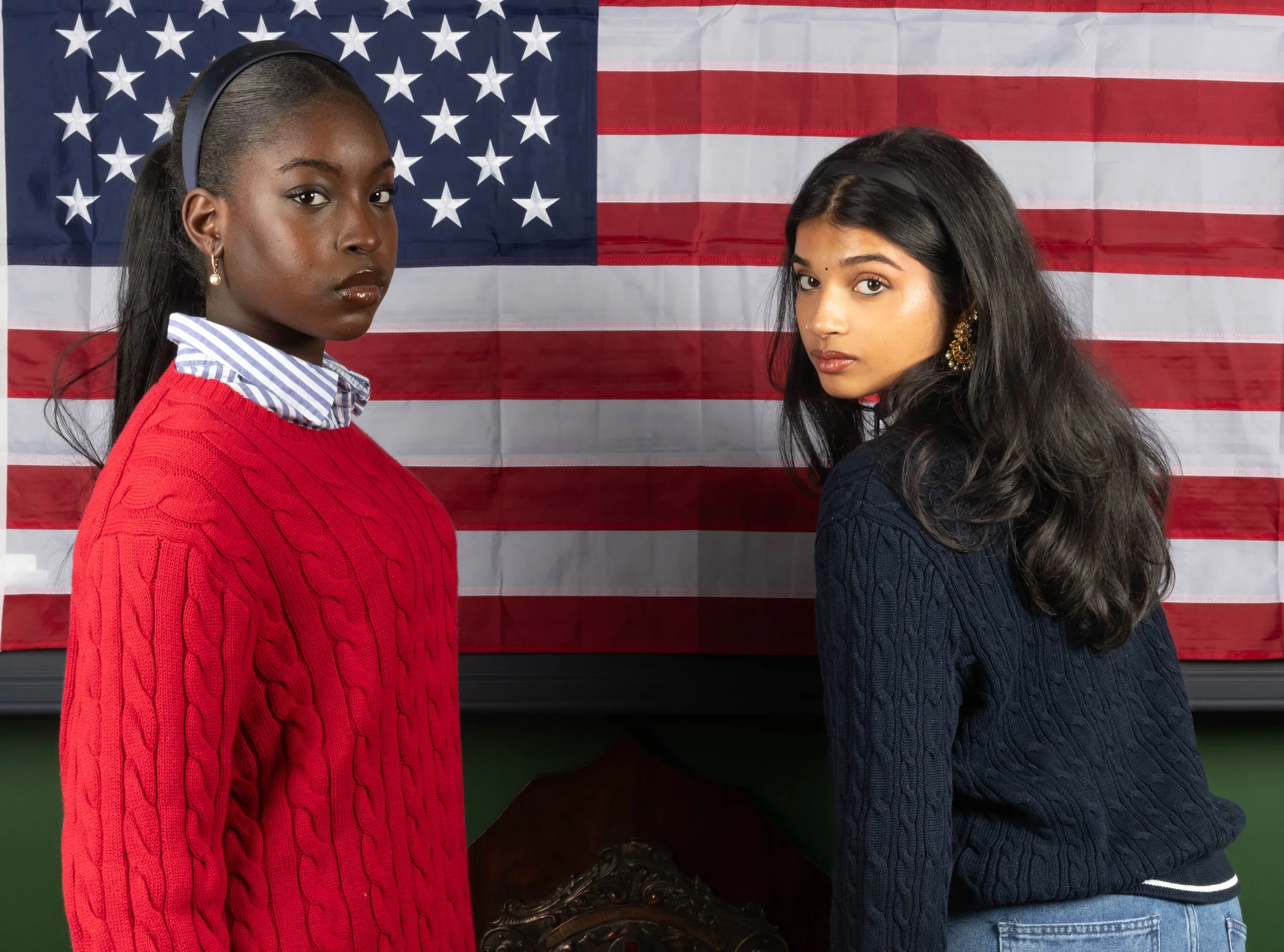 Two women stand side by side in front of an American flag, one wearing a red sweater and the other a navy sweater, both looking at the camera.