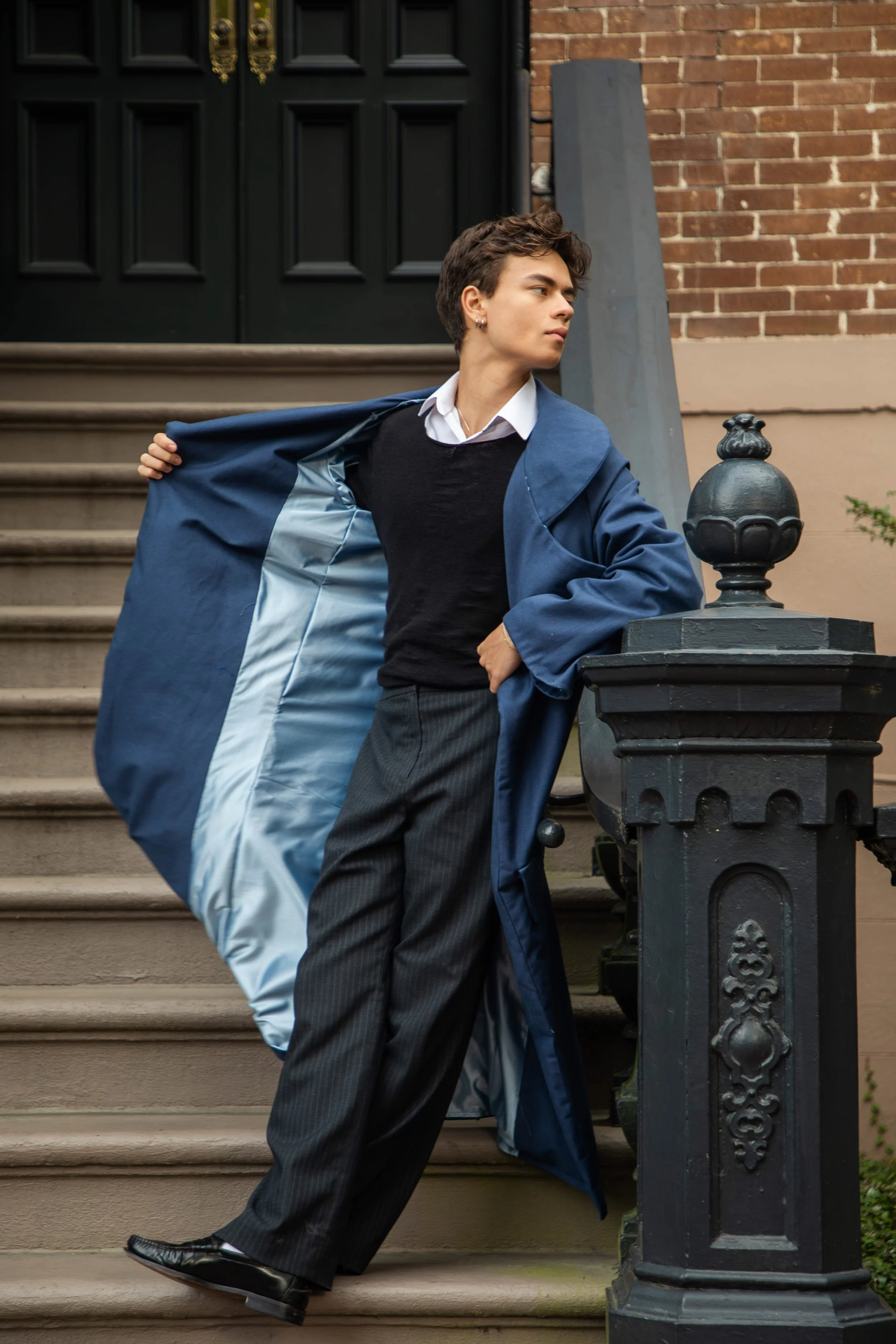 A young man in a white shirt, black vest, and pinstripe pants standing on stairs outside a brick building. He is adjusting his blue coat, looking to his left with a serious expression.