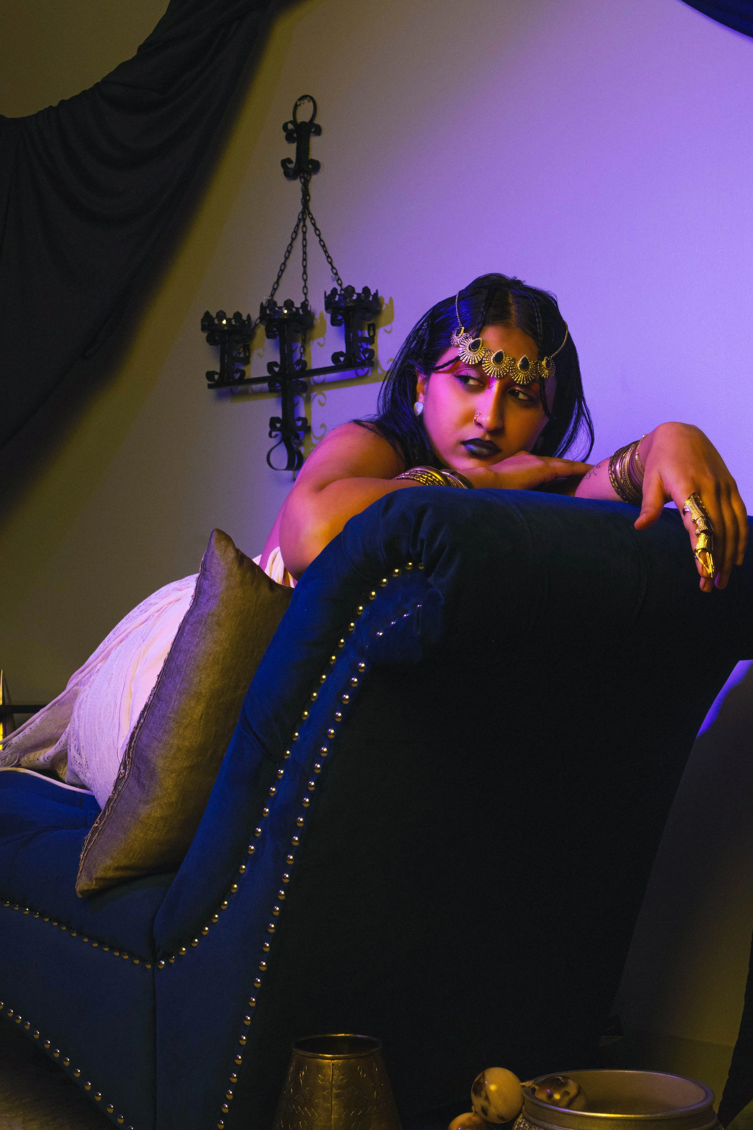 A woman with dark hair, wearing gold jewelry and dark lipstick, resting her arms on a black velvet furniture piece decorated with gold studs, in a room with dim purple and yellow lighting, decorative black candle holder wall piece, and a bowl of deco