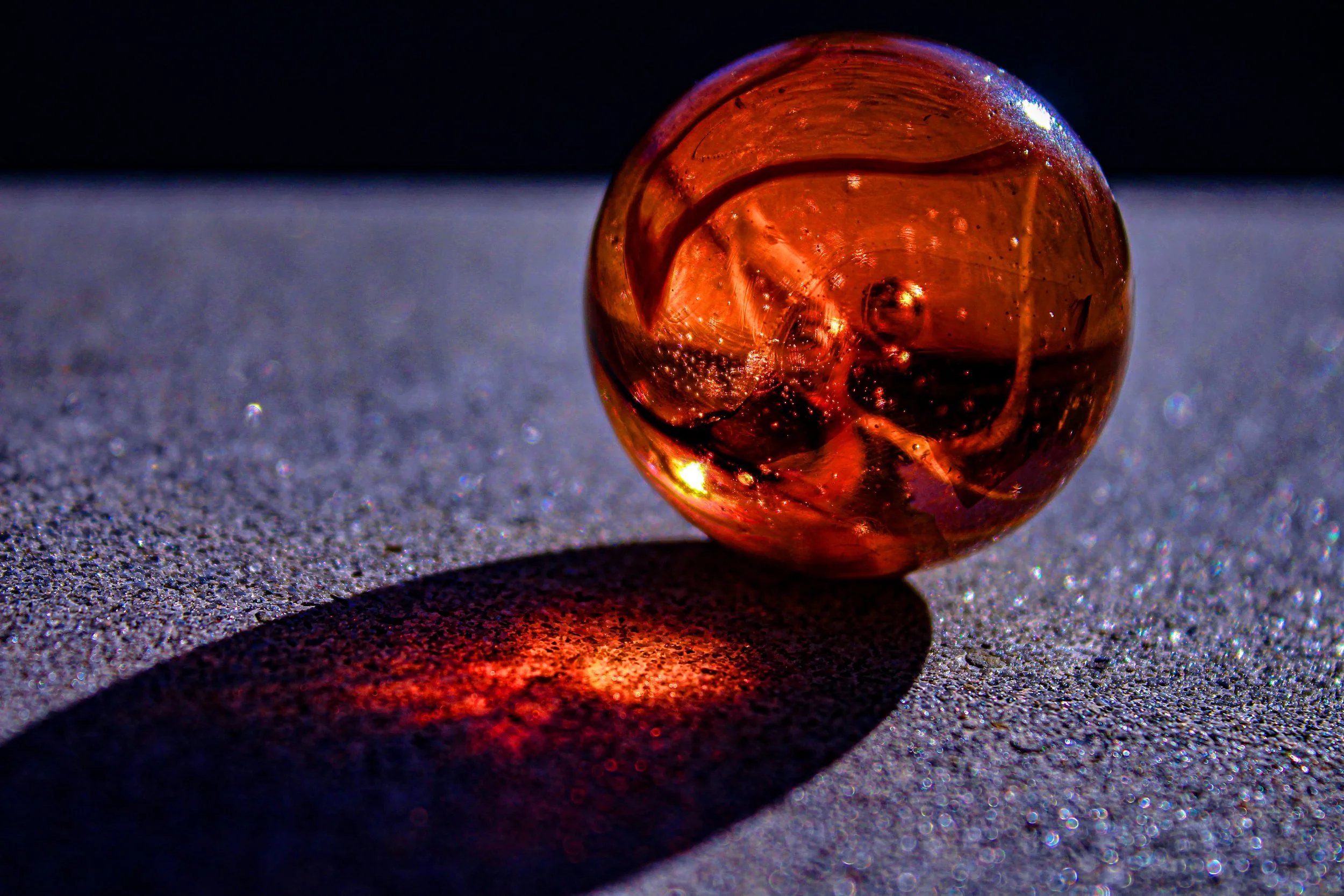 A small amber-colored glass marble on a sandy surface, casting a colorful shadow and reflection, with a dark background.