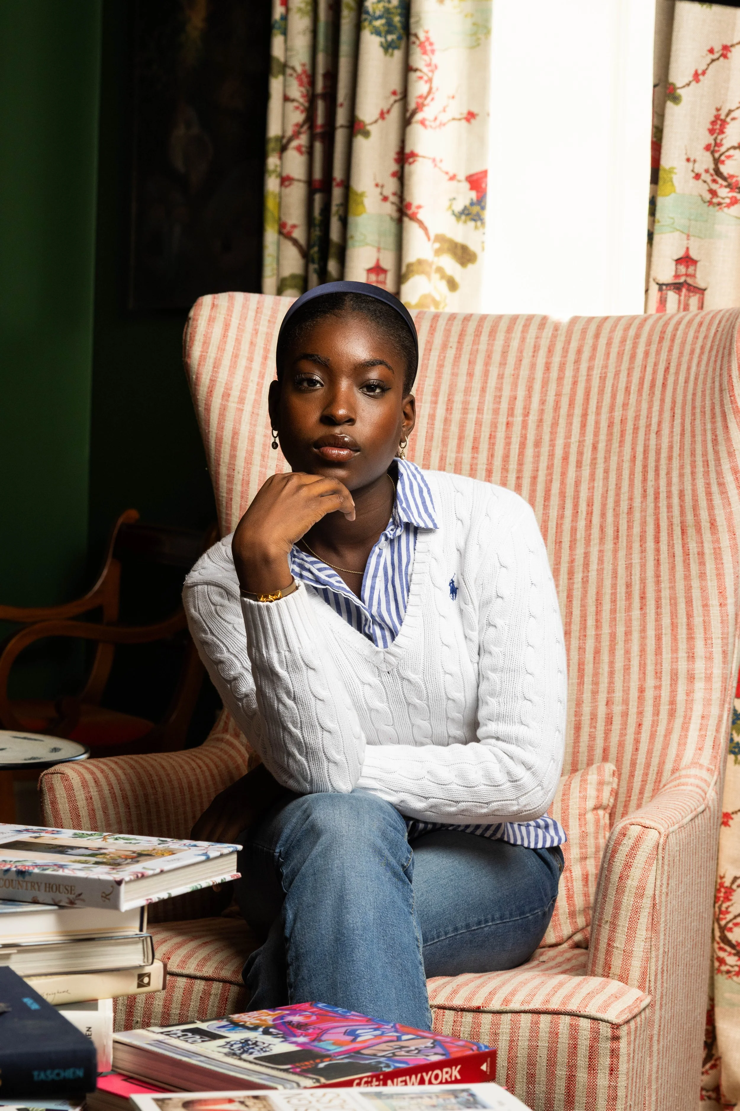 A young woman with dark skin and short hair sits on a pink and white striped armchair in a room with colorful patterned curtains and a window. She is wearing a white cable knit sweater over a blue and white striped shirt, with earrings and a gold bra