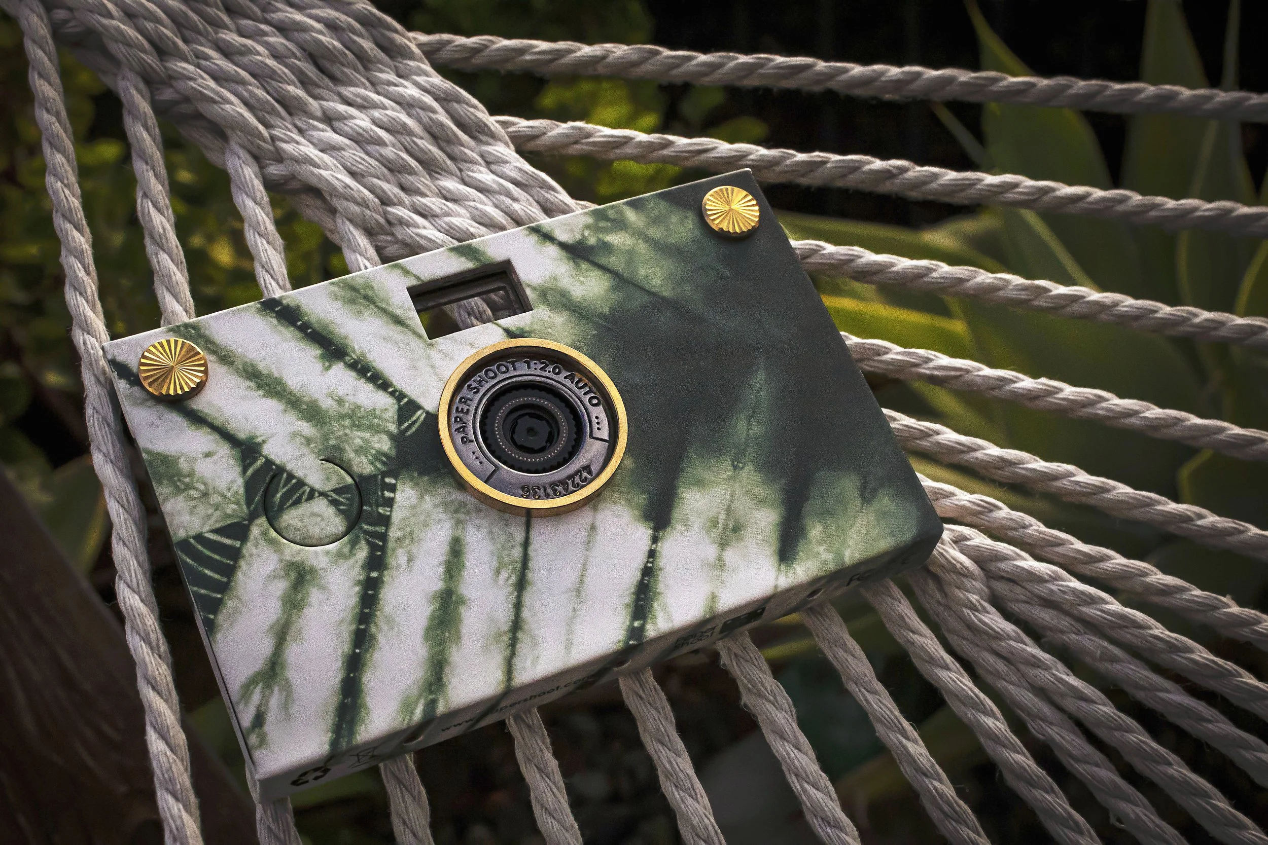 A camera box with green leaf and plant patterns resting on beige twisted rope with gold decorative accents in an outdoor setting.