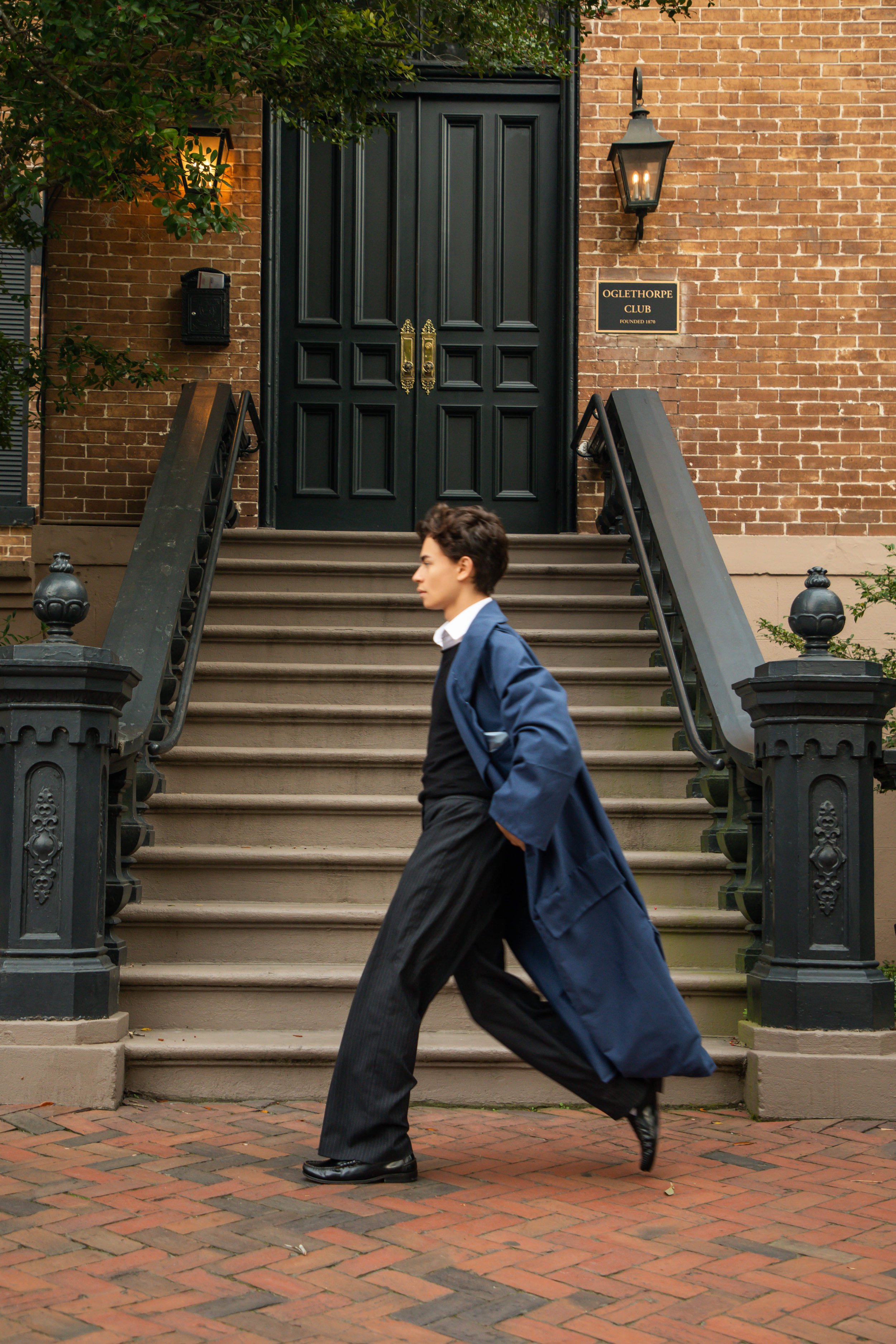 A person in a blue jacket and black pants walking past the stairs of a brick building with a black door, a wall-mounted lantern, and a brass plaque reading 'Oglethorpe Club Founded 1870'.