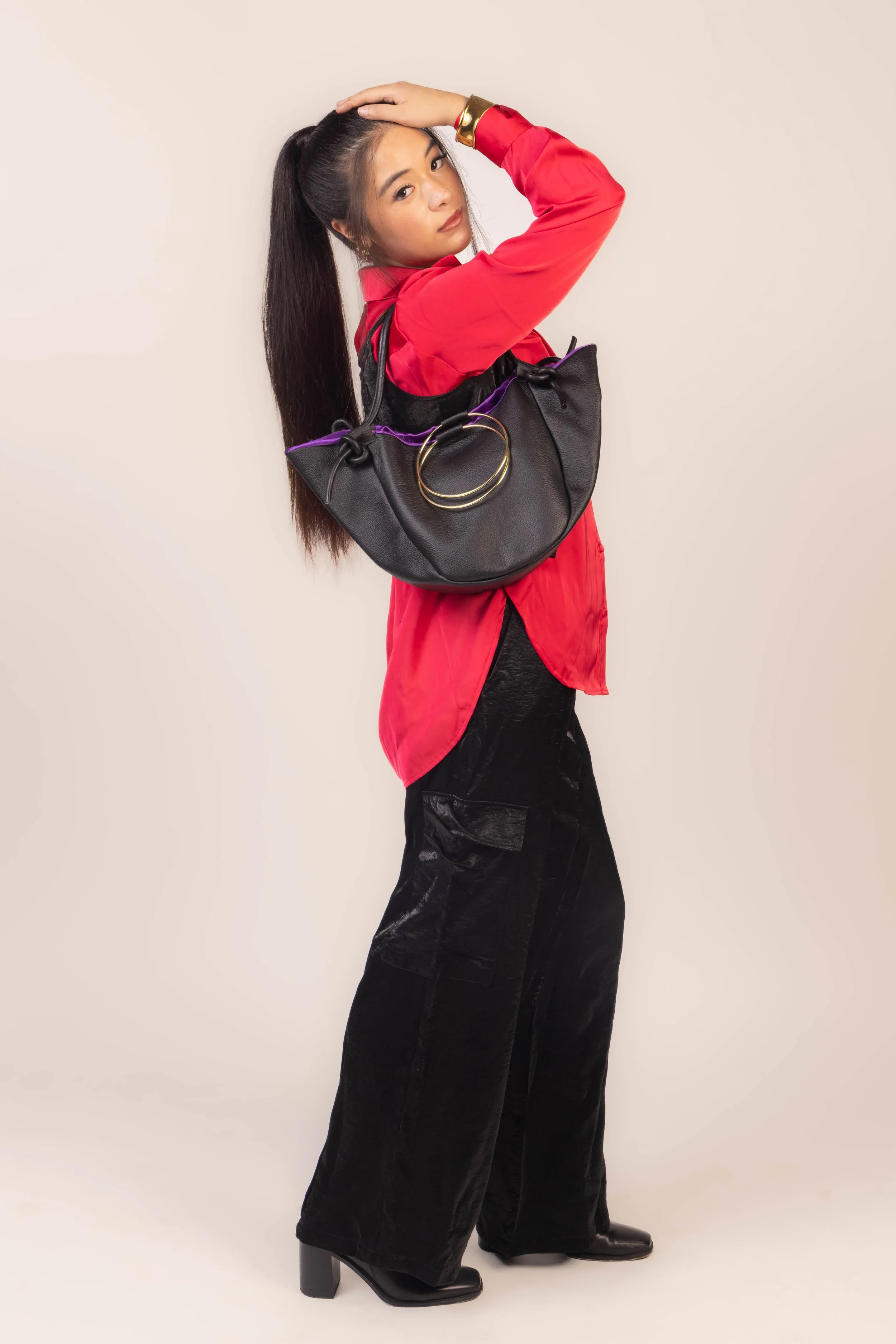 A woman with long dark hair in a high ponytail wears a red blouse with black fashionable wide-leg pants and black heeled boots. She has a large black handbag with purple accents and gold hardware draped over her shoulder. She poses against a plain, l
