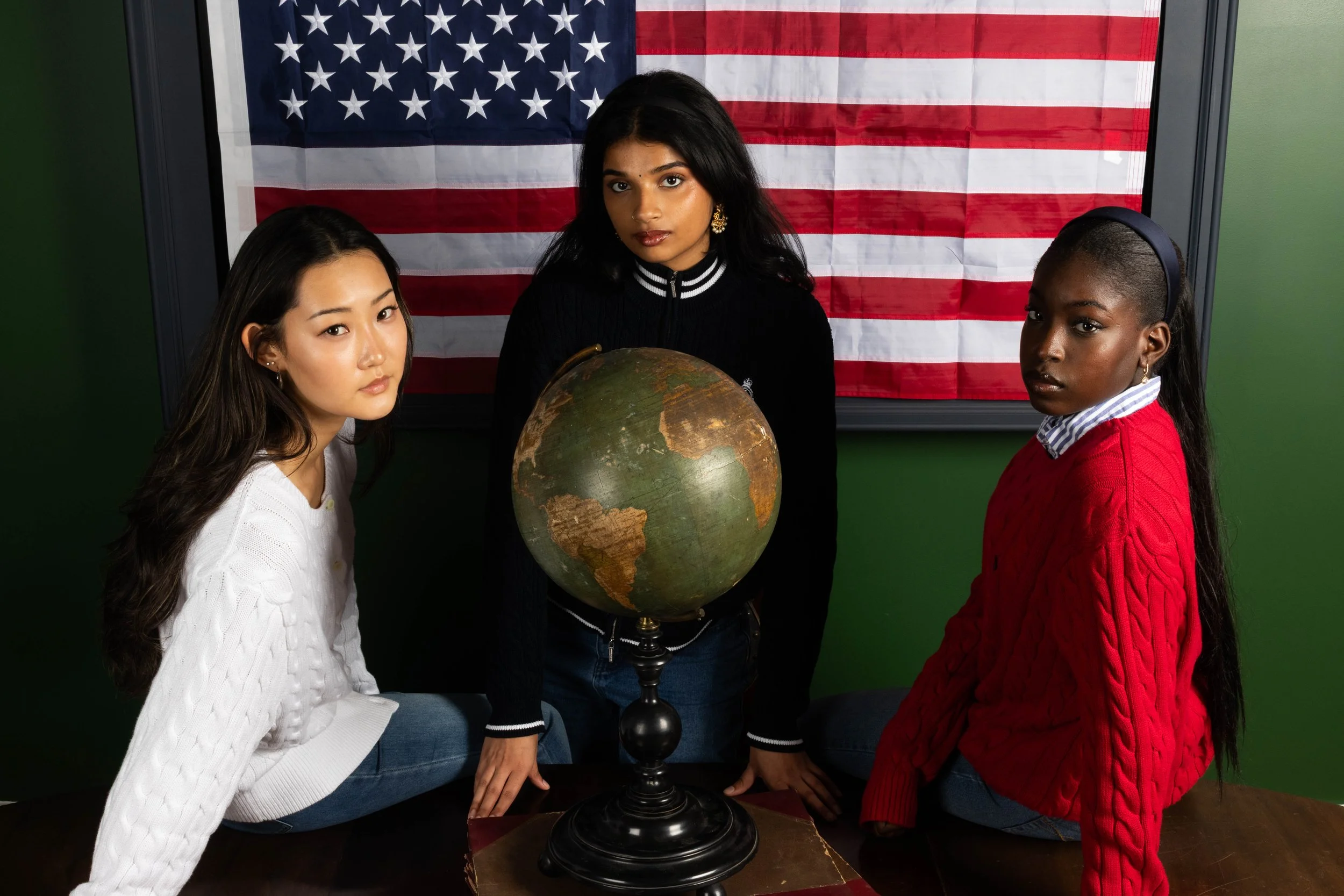 Three women are gathered around a globe on a table with an American flag backdrop. One woman is wearing a white sweater, another is wearing a black jacket with white stripes, and the third is wearing a red sweater with a collared shirt underneath.