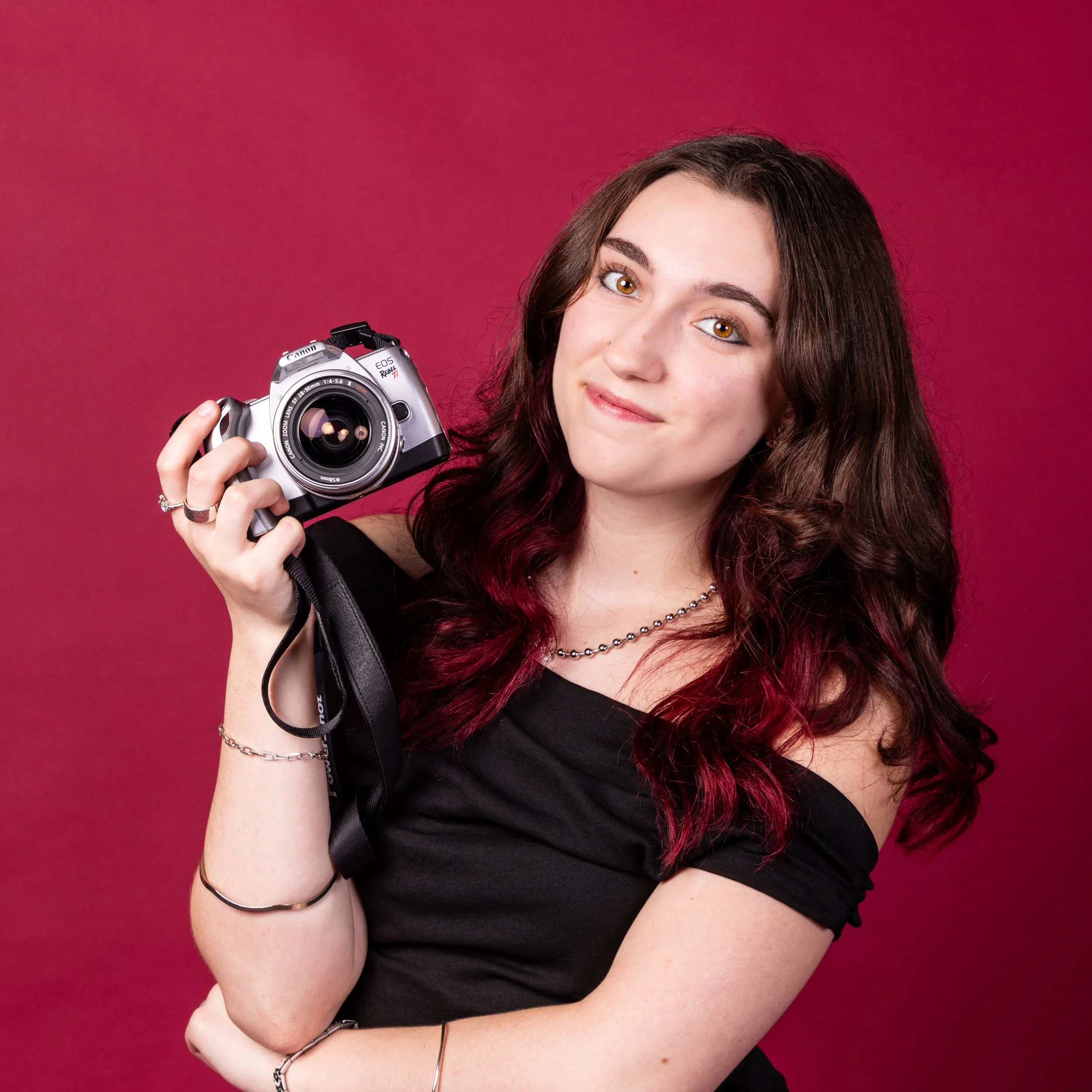 A young woman with brown and red hair, brown eyes, and fair skin, smiling slightly, holding a camera in her right hand, standing against a red background.