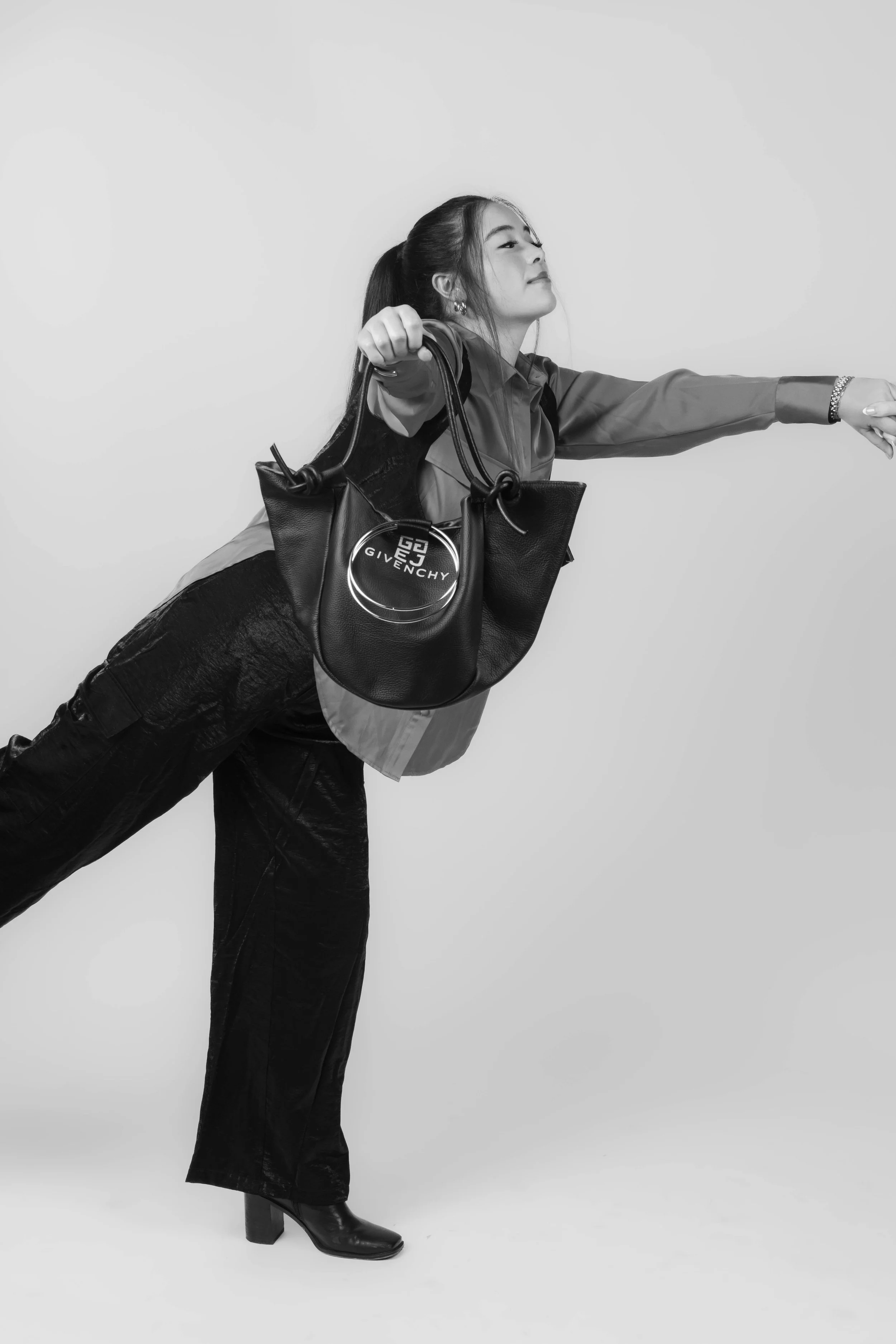 A woman in black boots and dark pants is stretching her arms forward while holding a handbag with the Givenchy logo, wearing a jacket, against a plain light background.