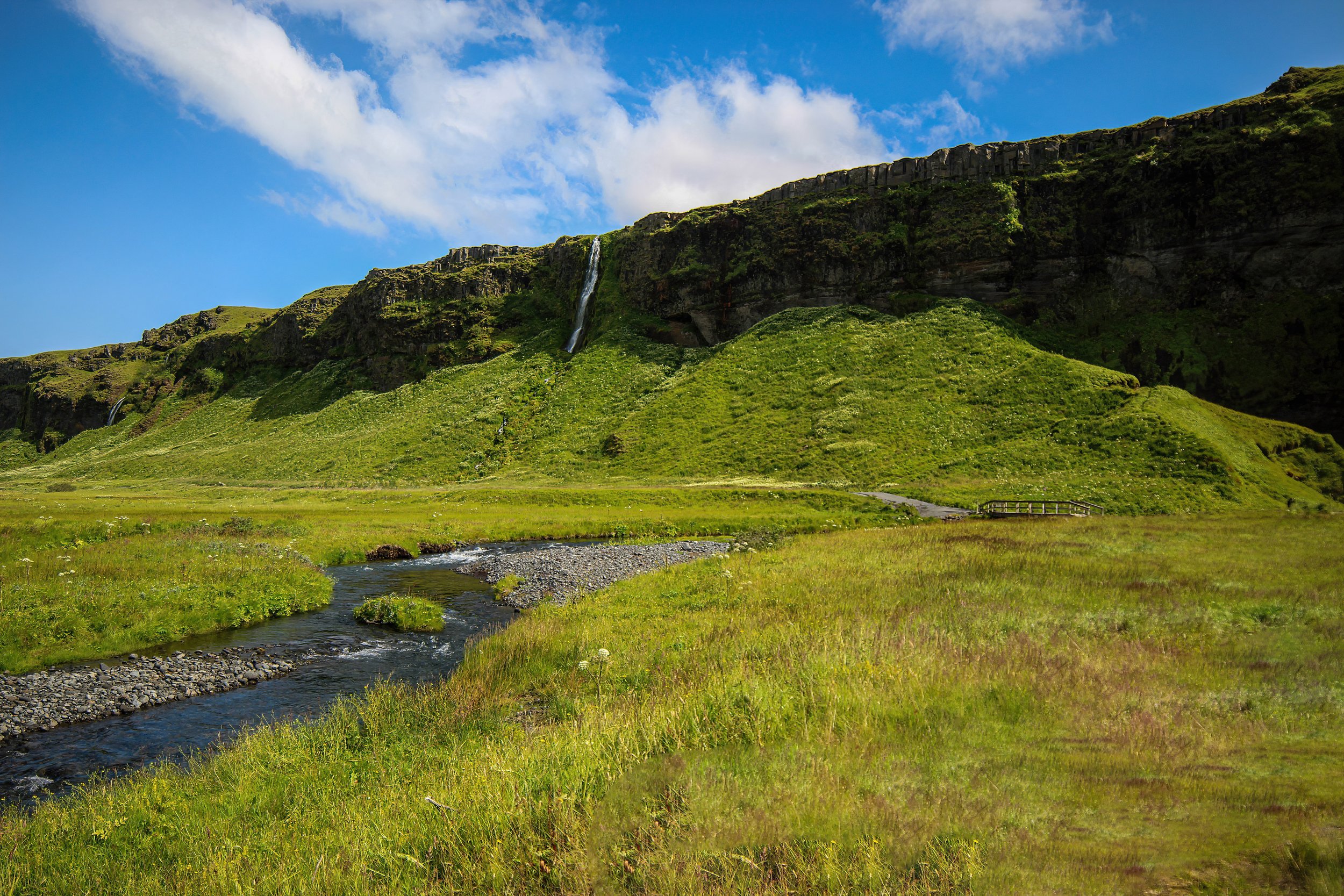 A green hillside with a waterfall and a small stream in the foreground, under a partly cloudy blue sky.