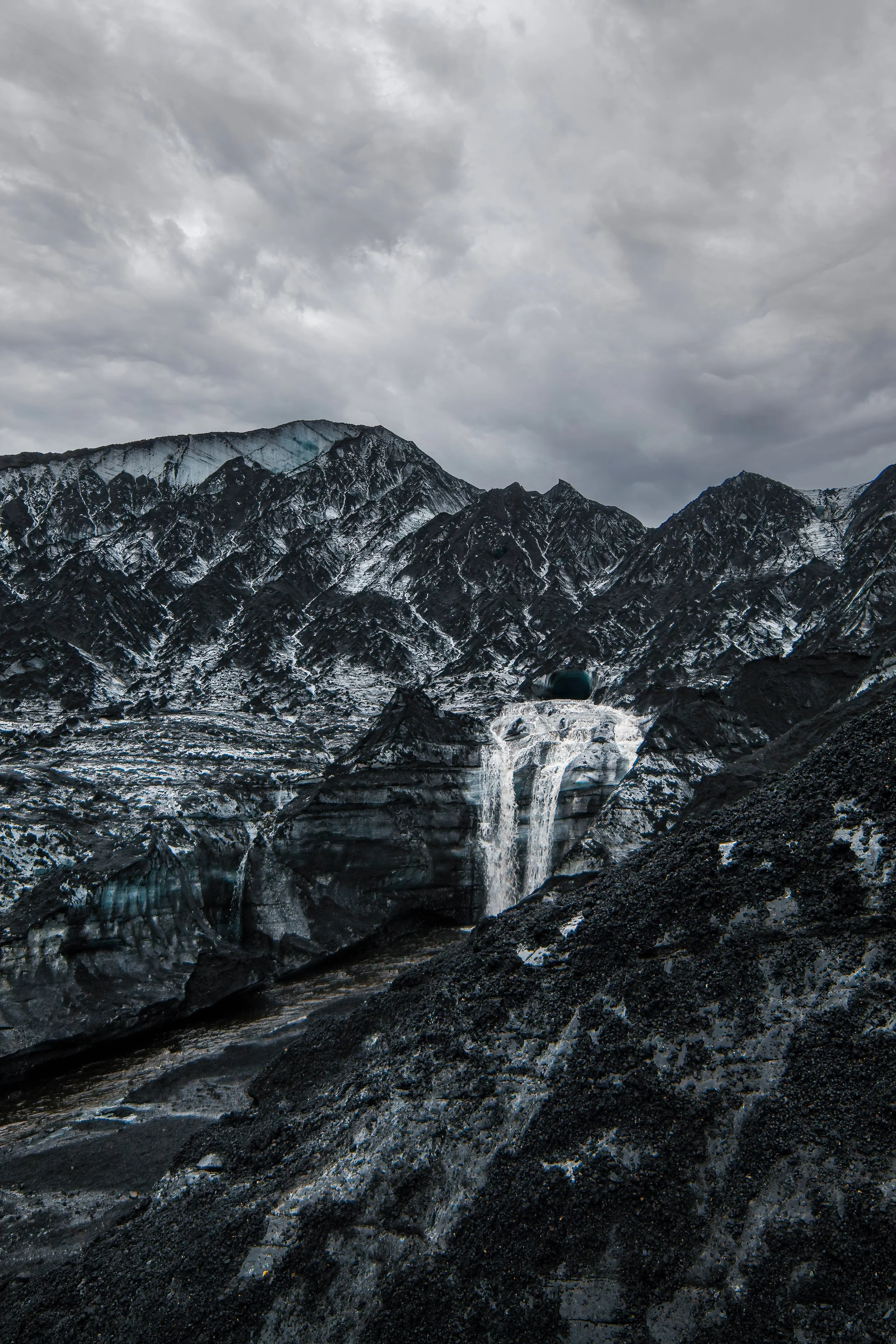 Snow-capped mountains under a cloudy sky with a waterfall cascading down a dark rocky terrain.