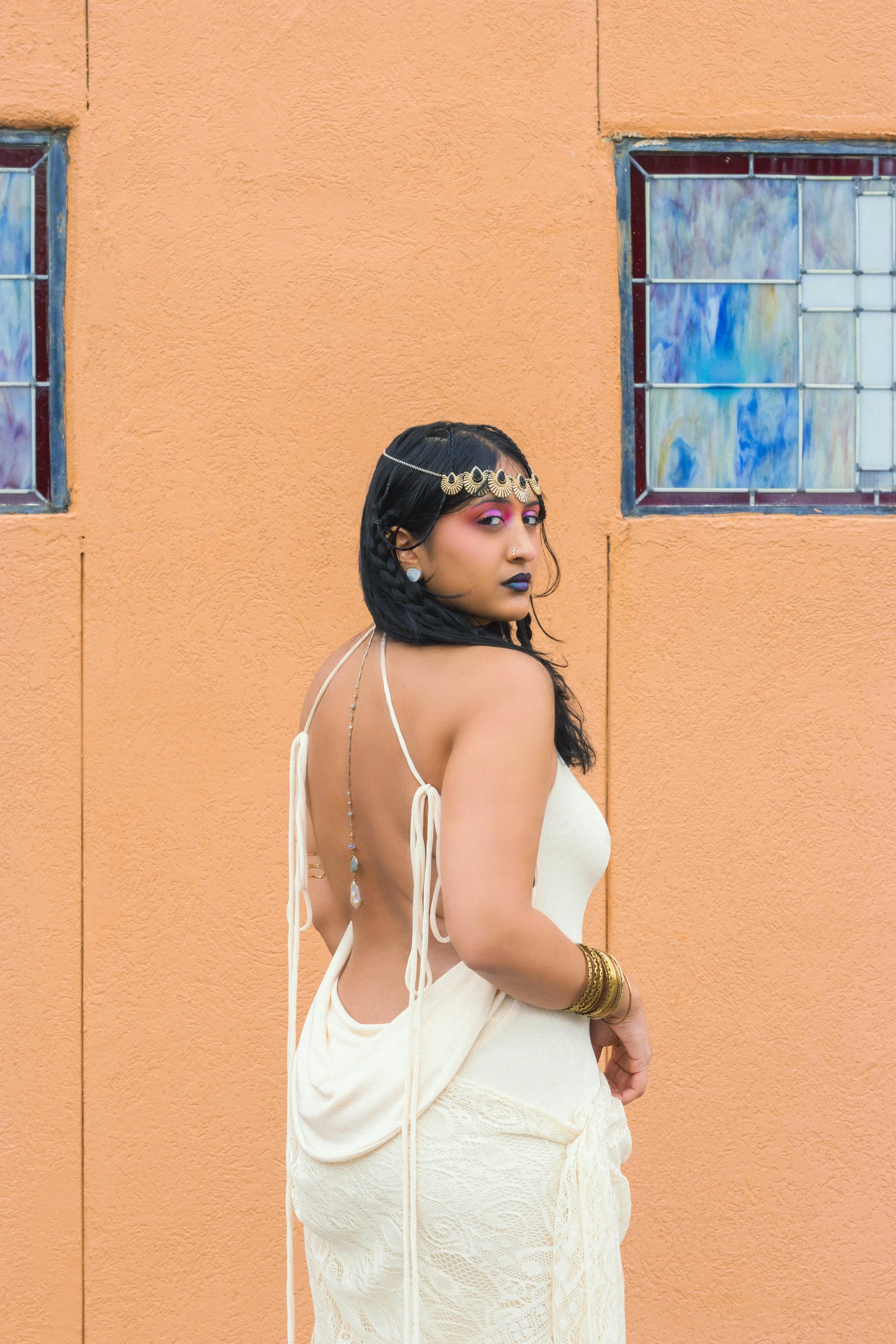 A woman with dark hair styled in braids and adorned with a gold headpiece, wearing bold makeup with purple eyeshadow and dark lipstick, dressed in a cream-colored outfit with backless design, standing in front of an orange wall with stained glass win