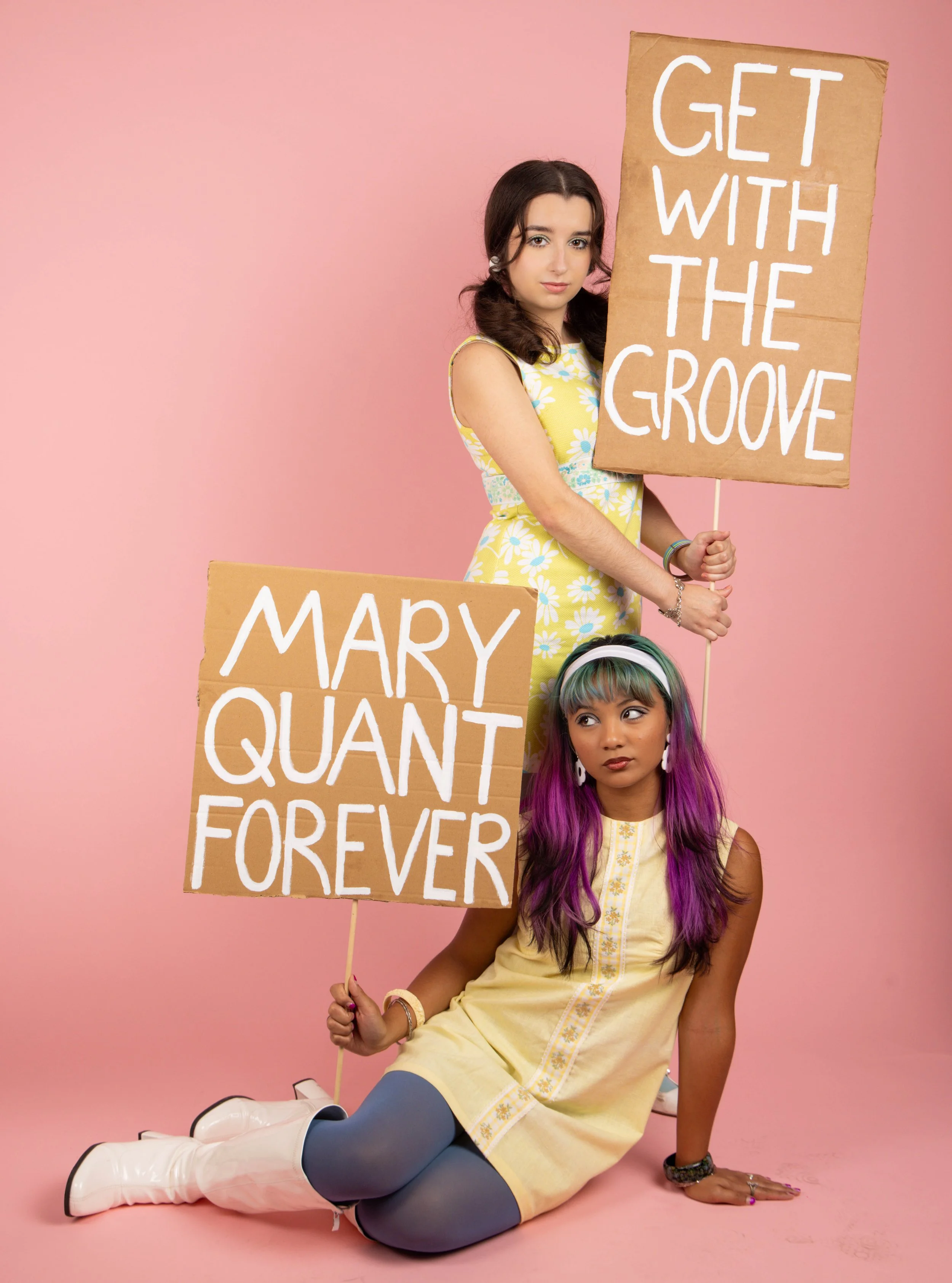 A young woman with brown hair sitting on the floor against a pink background, holding a cardboard sign that reads 'GET WITH THE GOOSE'. She wears a sleeveless yellow dress with flower patterns, blue tights, and white boots. Another person, partially 