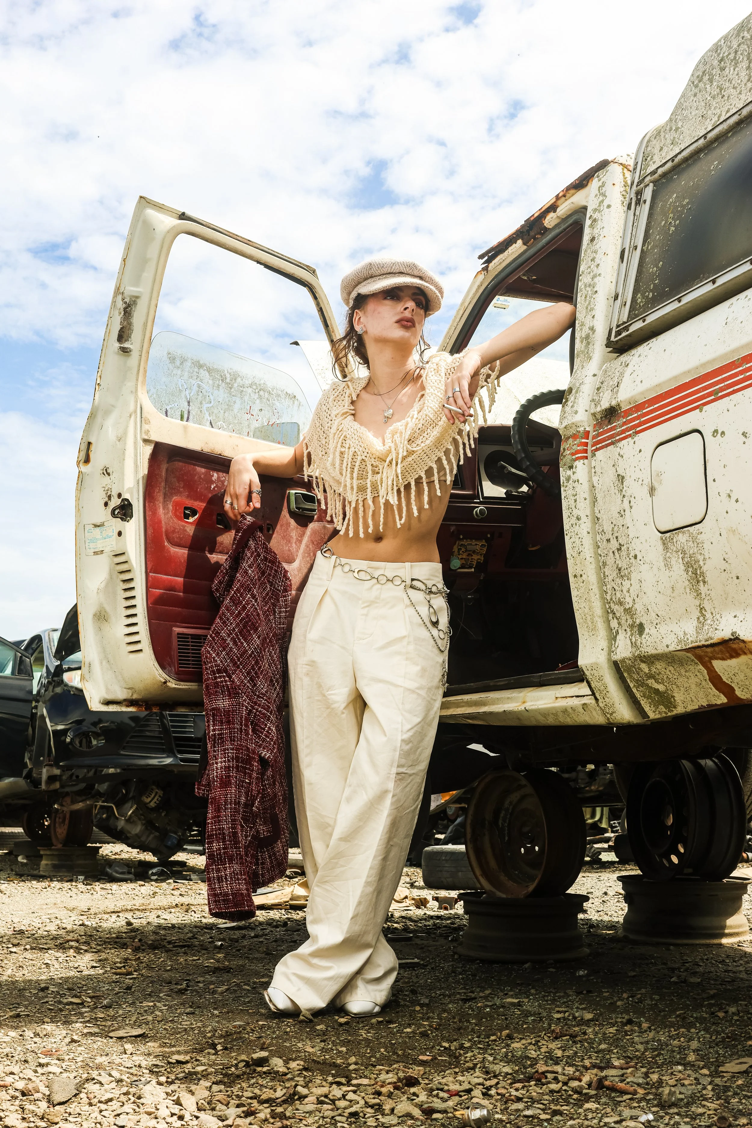 A woman standing in a junkyard beside an old, rusty vehicle. She is wearing a knit top, white pants, and a beige hat. The sky is partly cloudy.