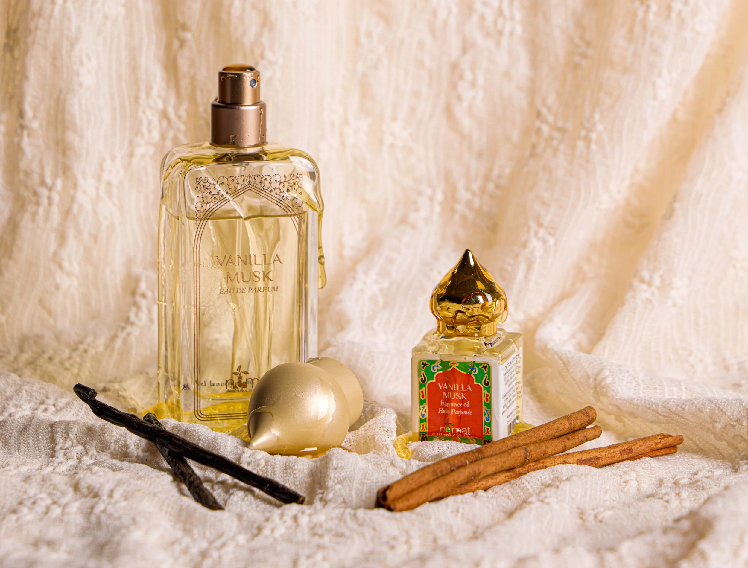 A decorative display of vanilla musk perfume, vanilla fragrance oil, a vanilla bean, cinnamon sticks, and a small ornamental object on a textured fabric background.