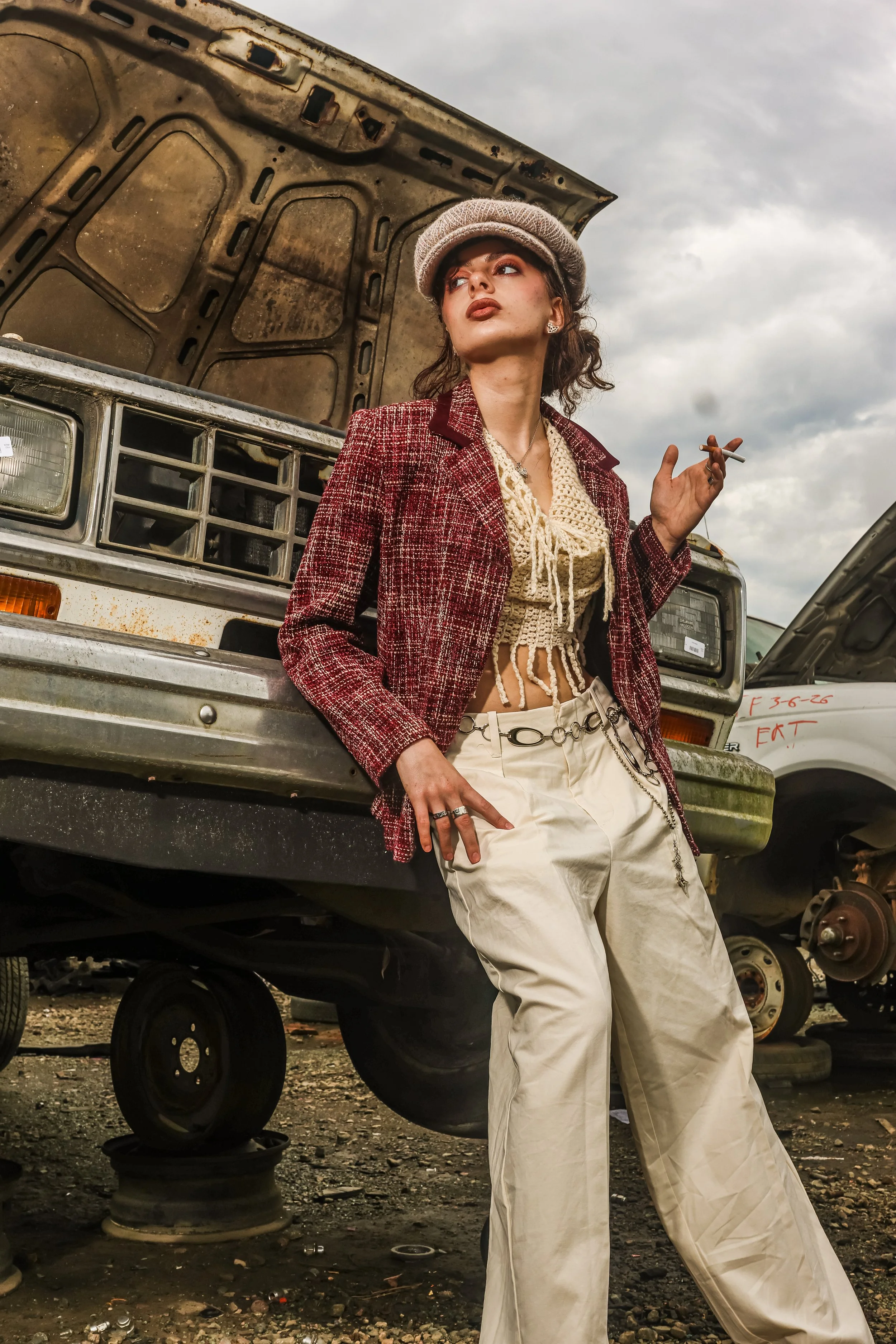 A young woman with makeup and curly hair, wearing a beret, a patterned blazer, a crochet top, and baggy pants, stands in a scrapyard next to a rusty, open hood car. She is holding a cigarette and looking off into the distance.