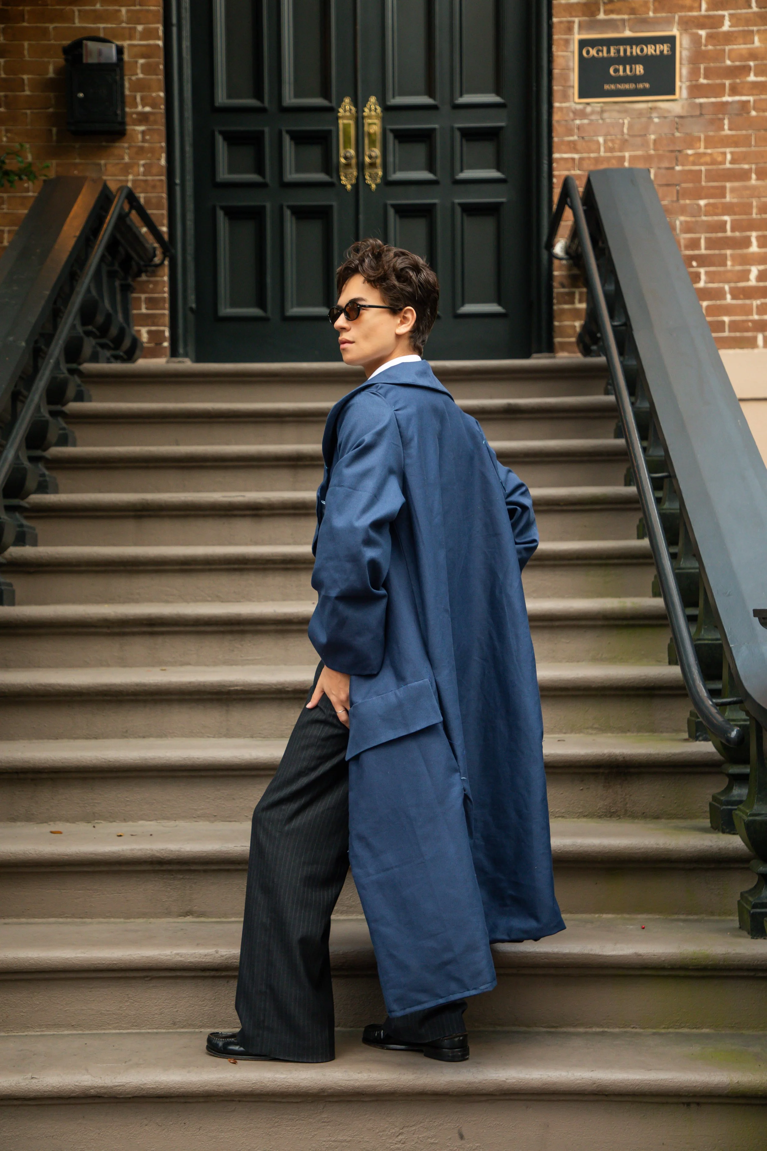A young man with curly hair wearing sunglasses, a blue trench coat, and black pinstripe pants walking down brownstone stairs in front of a brick building with a black door and a sign that reads 'Oglethorpe Club'.