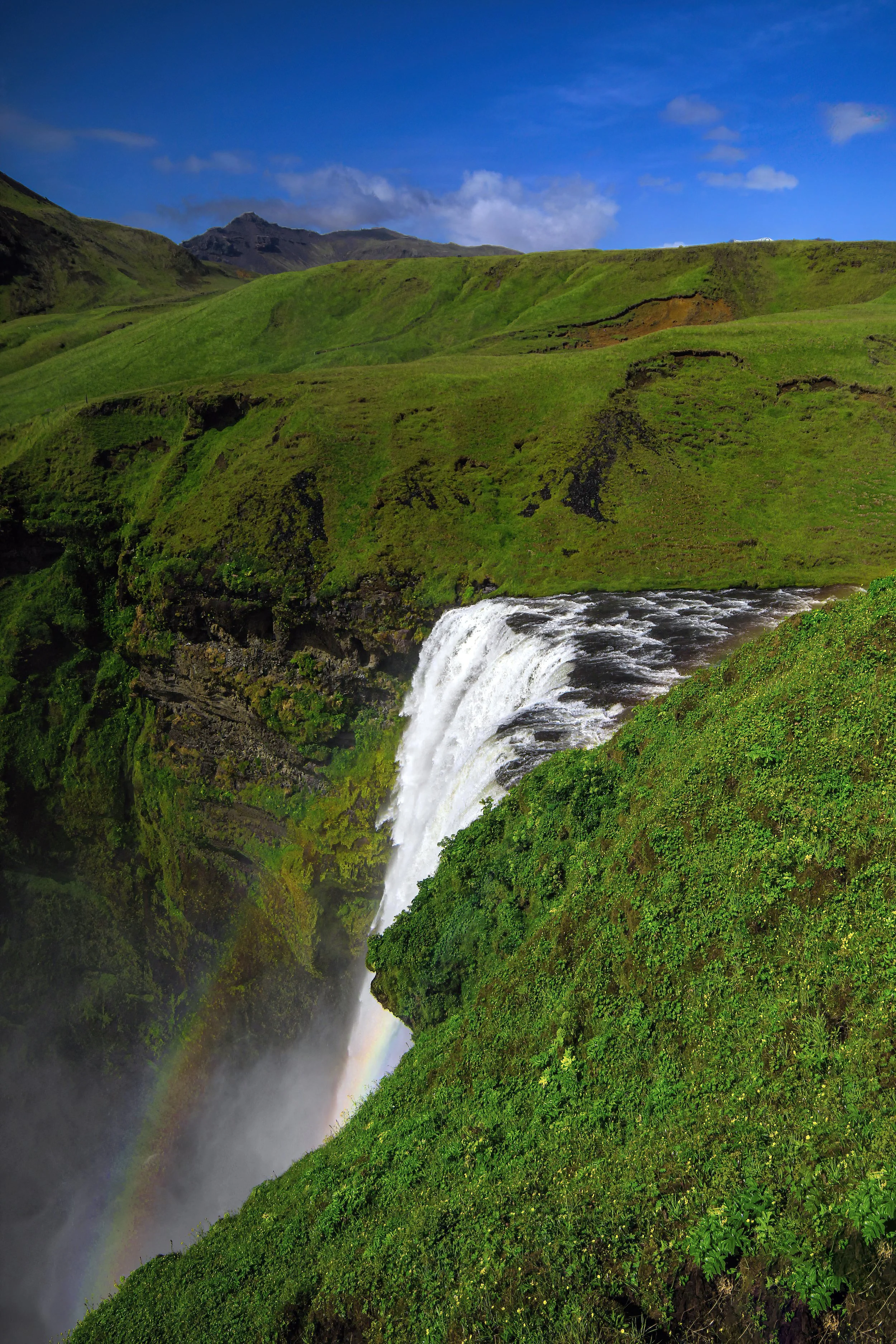 A scenic view of a waterfall cascading down green cliffs with a rainbow at the bottom, green hills, and a bright blue sky with some clouds.