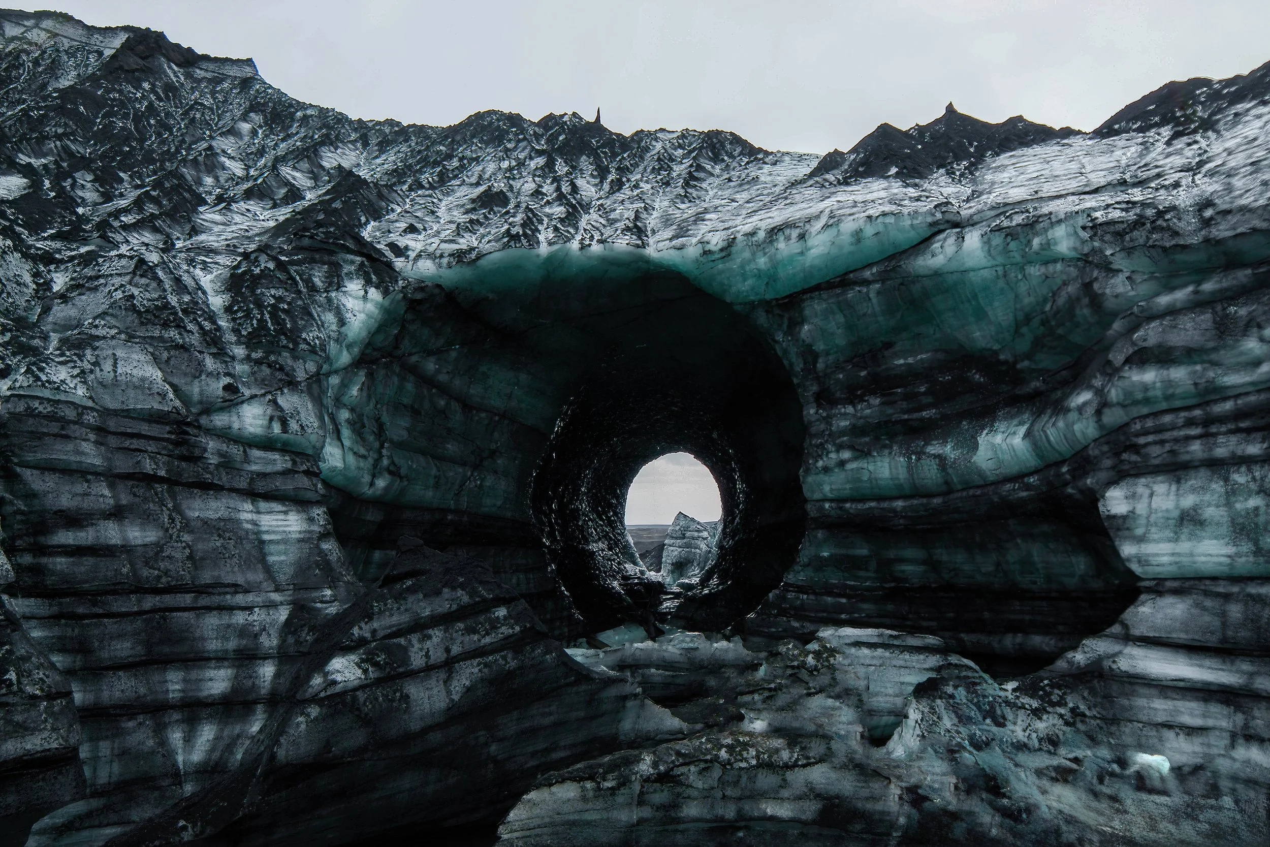 A glacier with a natural ice arch and a tunnel through the ice, near a mountain landscape with snow-capped peaks.