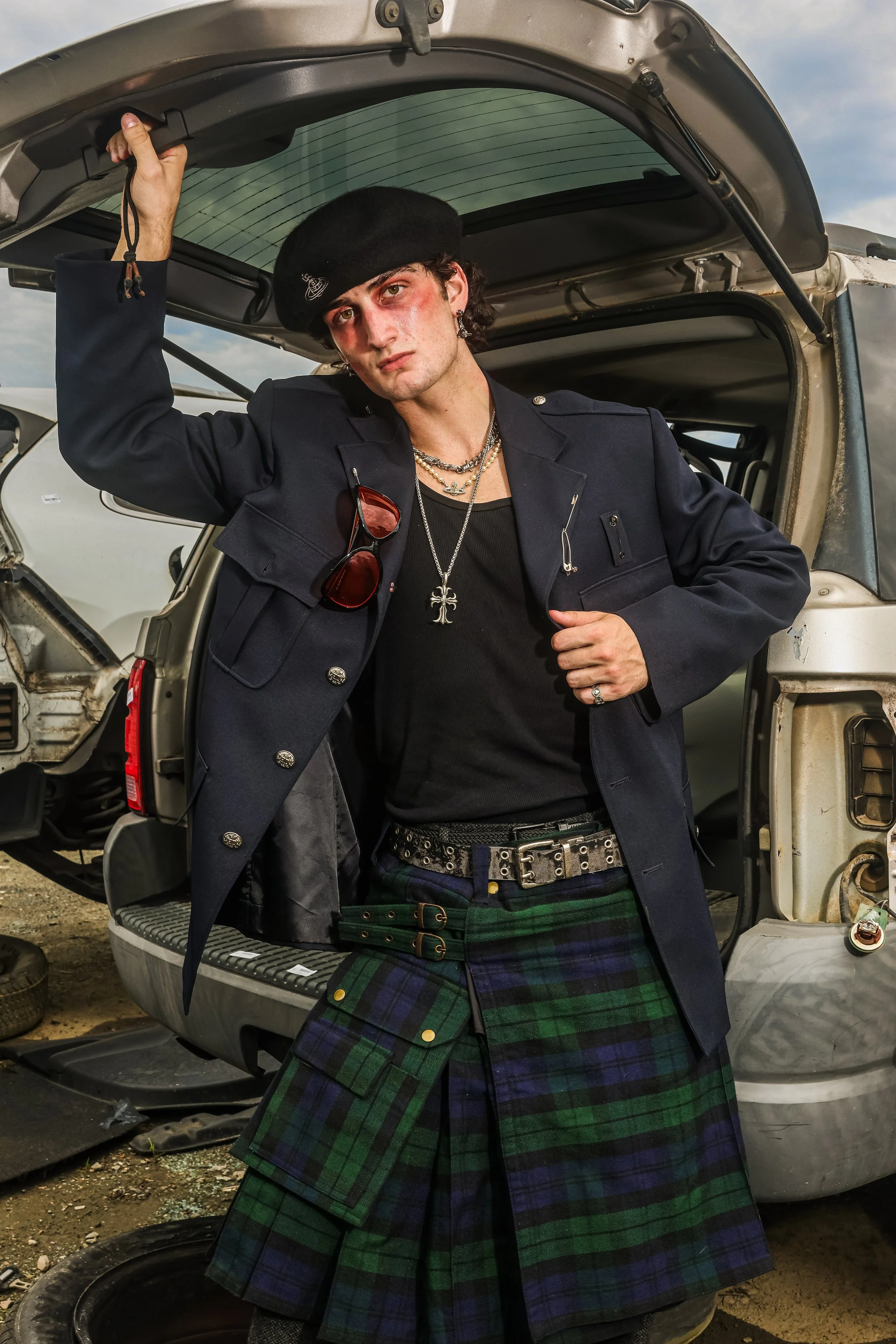 Young man with makeup and jewelry standing in front of a damaged vehicle. He is wearing a black beret, a dark blazer, sunglasses hanging from the blazer, a black shirt, a cross necklace, and a green and blue tartan kilt. His hand is holding the edge 