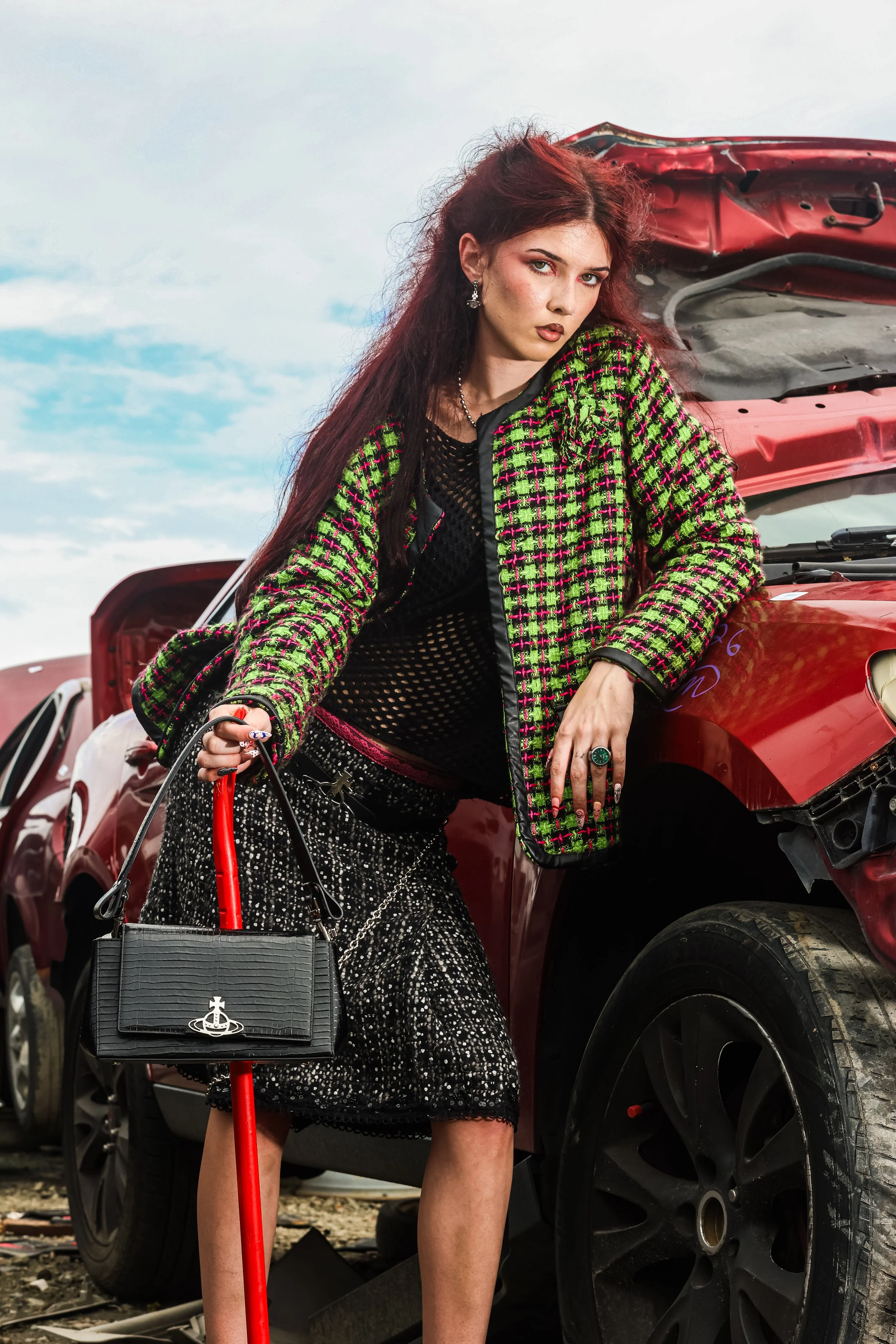 A woman with red hair stands in front of a damaged red car. She is wearing a black and green patterned jacket, a black netted top, and black and white patterned pants. She holds a black handbag with a red cane. The sky is cloudy.