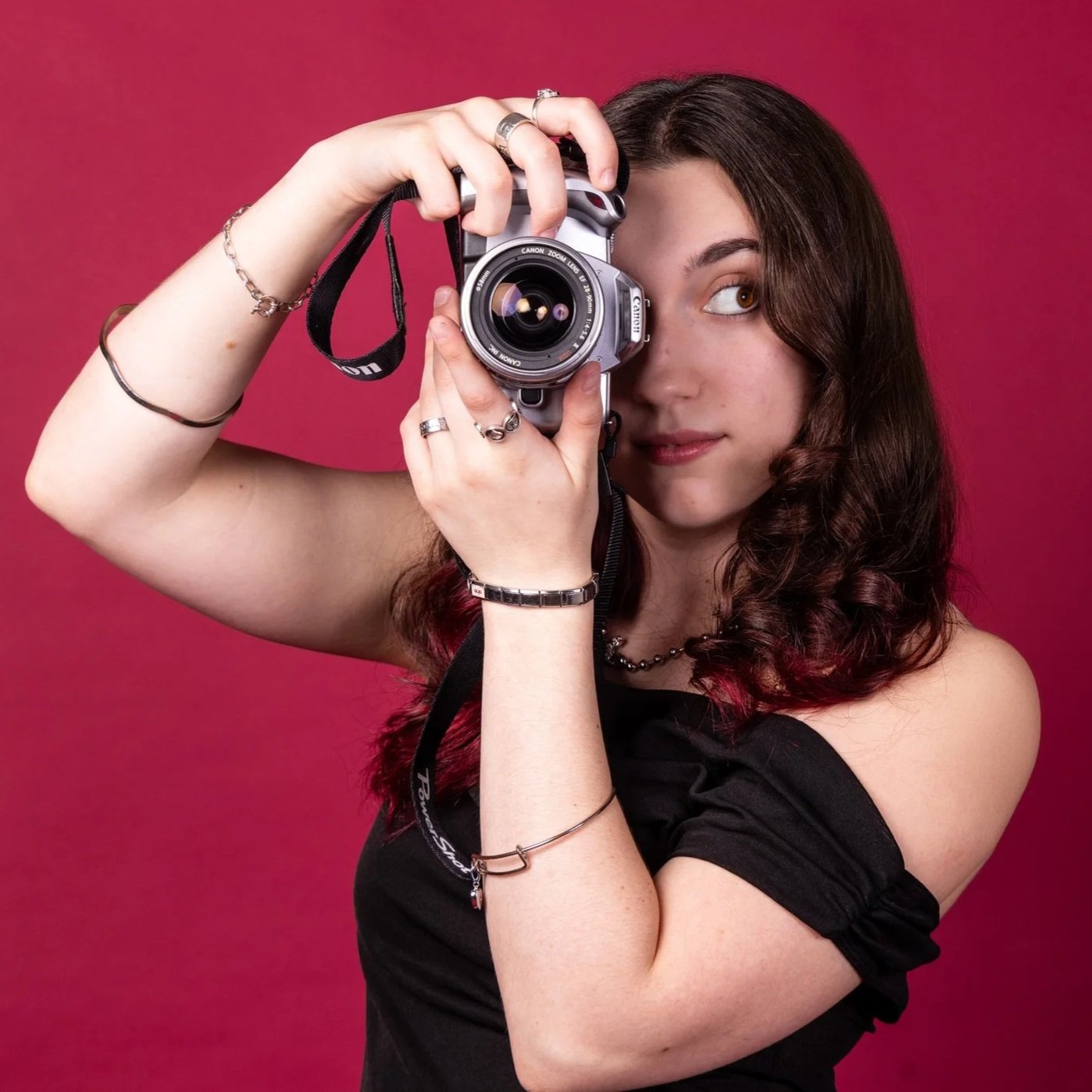 A young woman with wavy brown hair and brown eyes holding a Canon camera, taking a photo against a pink background.