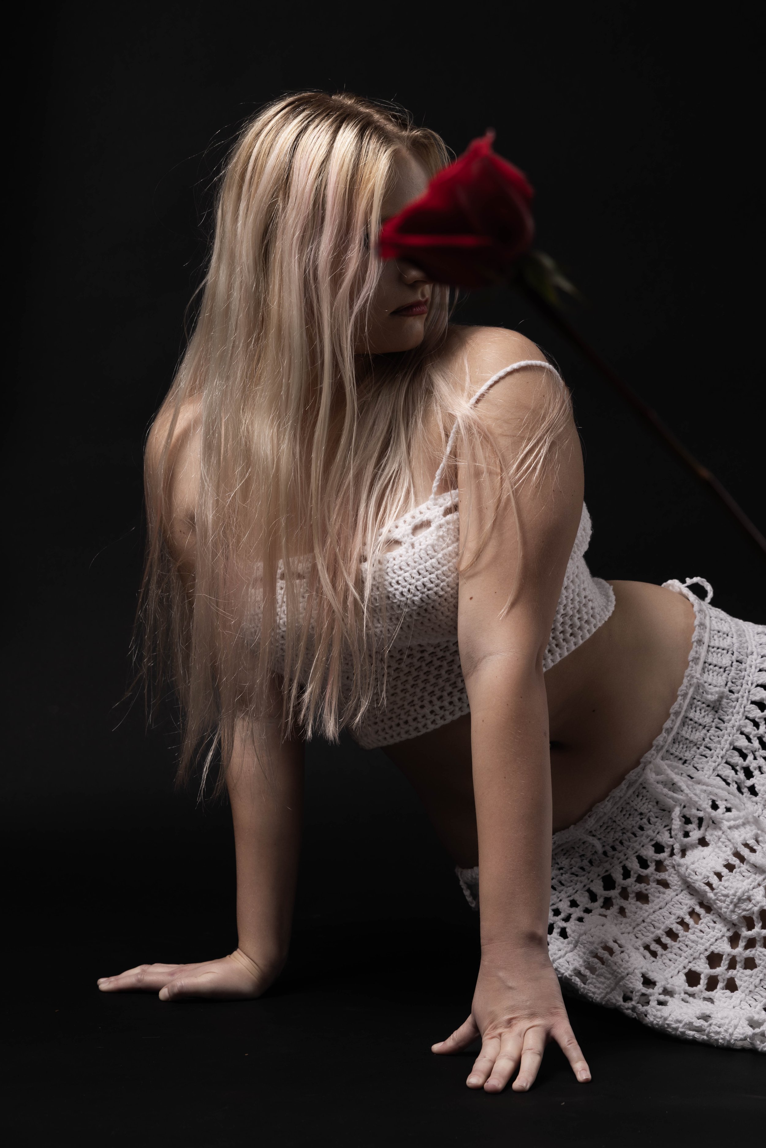 A woman with long blonde hair wearing a white crocheted top and shorts is in a pose on her hands and knees against a black background. Her face is obscured by a red rose she is holding close to her face.