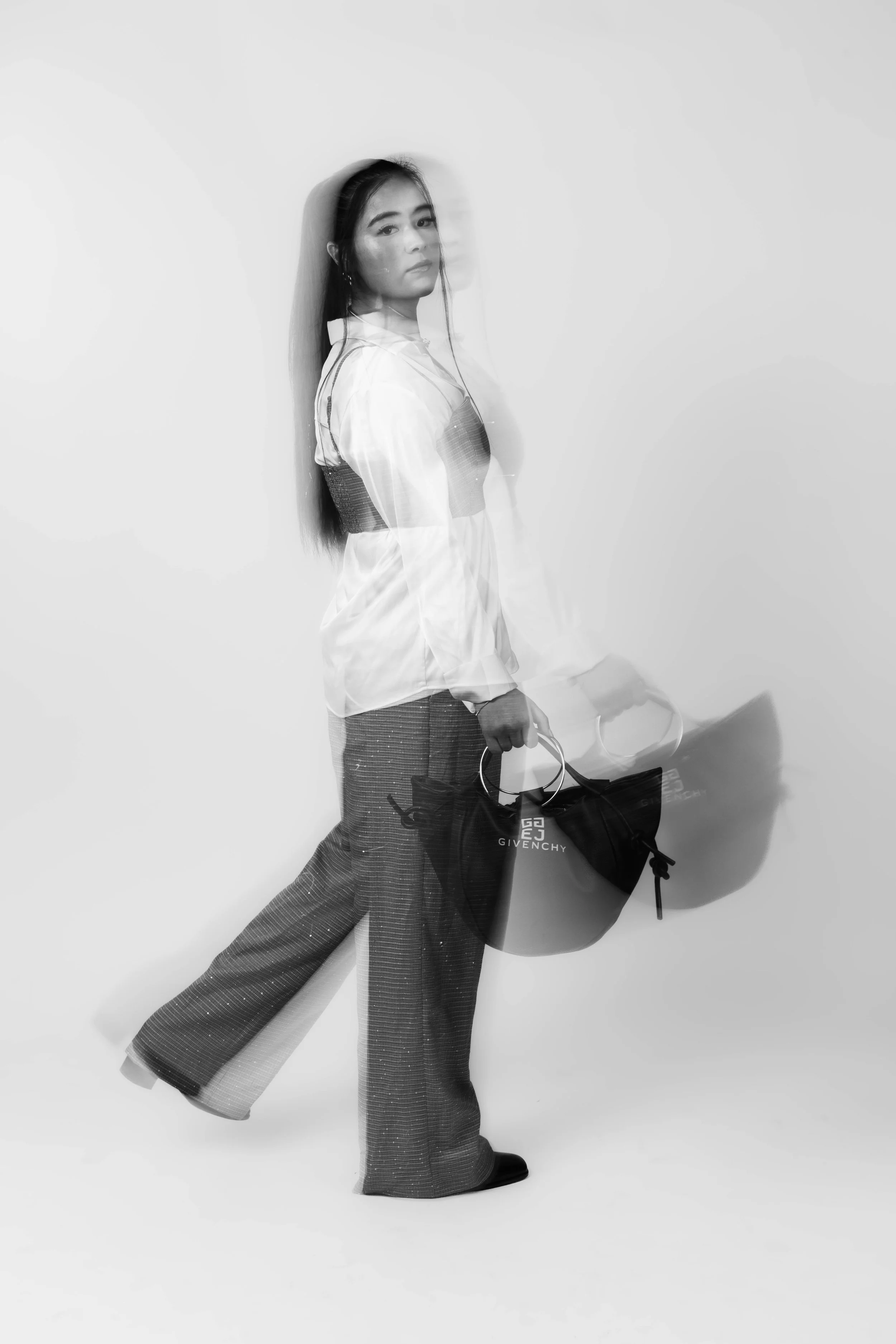 Black and white photo of a woman with long dark hair, standing against a plain light background. She wears a white blouse, patterned wide-leg pants, and black shoes. She holds a shopping bag with a Givenchy logo and a pair of sunglasses, creating a layered, motion-blur effect.
