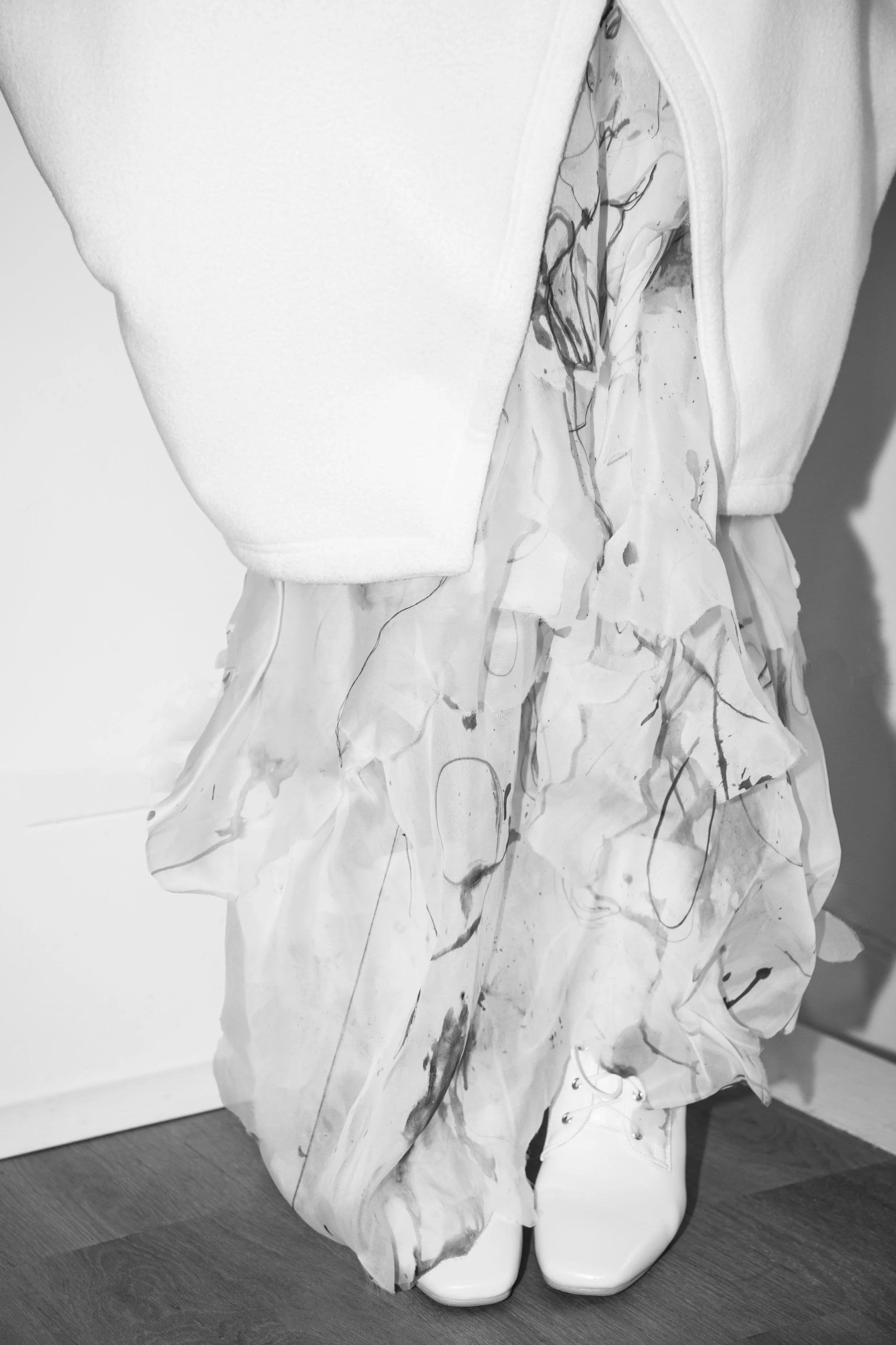Close-up of a person wearing a white, long-sleeve top, an abstract patterned skirt, and white shoes, standing on a wooden floor.