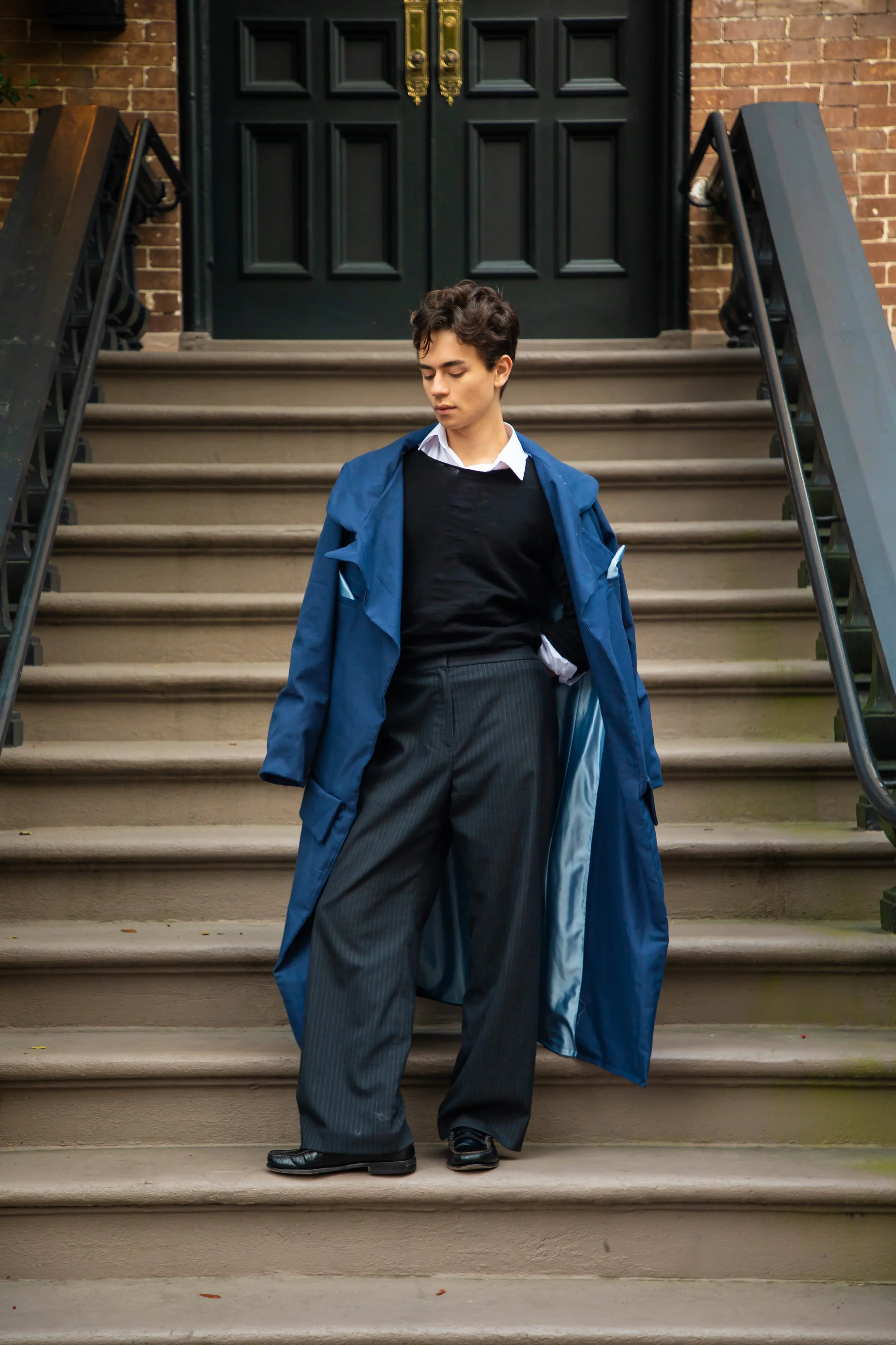 Young man with dark hair standing on beige steps in front of a black door, wearing a dark sweater over a collared white shirt, gray pinstripe pants, and a blue coat draped over his shoulders, looking down with a serious expression.