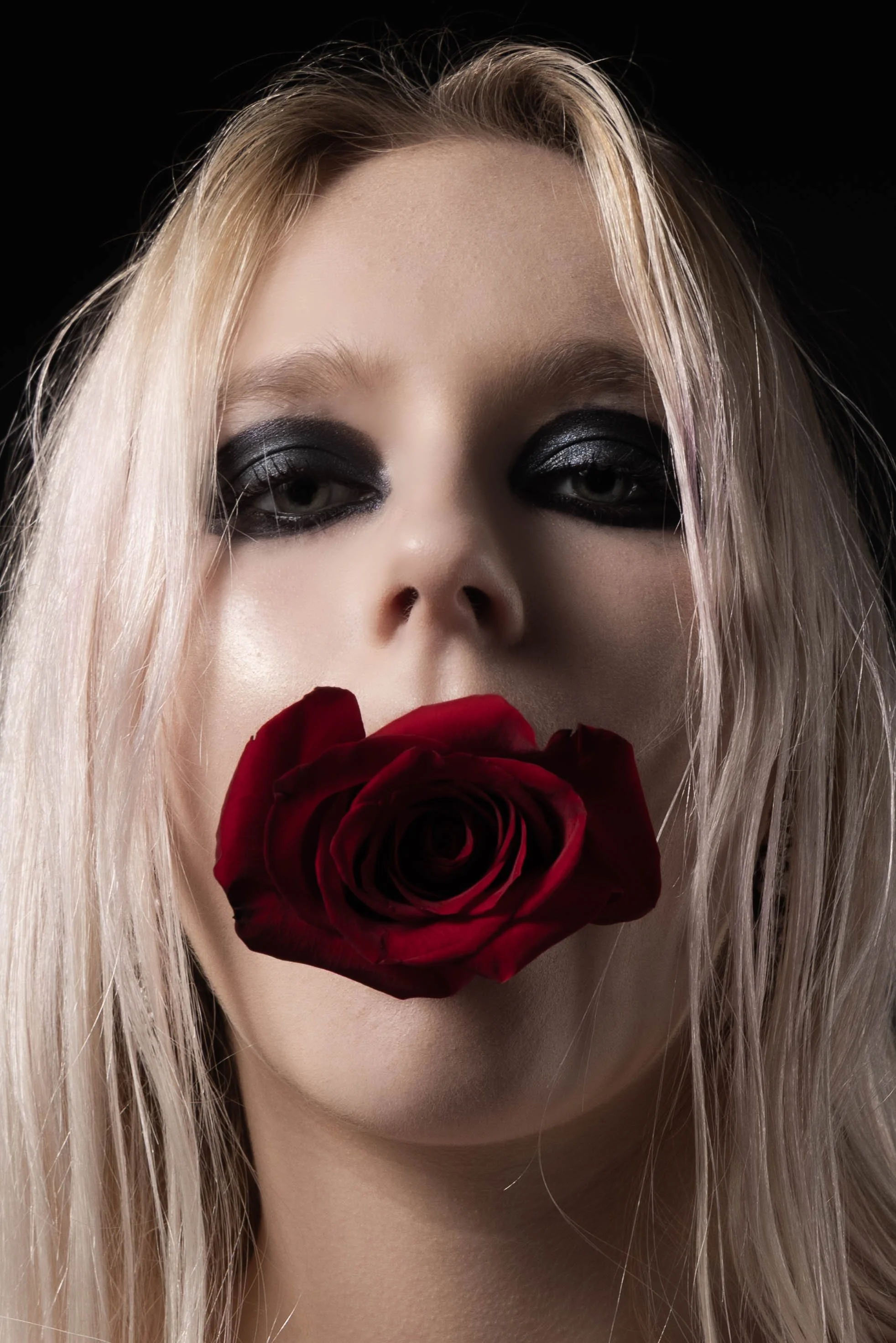 A woman with platinum blonde hair and dark makeup, holding a red rose in front of her lips, close-up portrait.