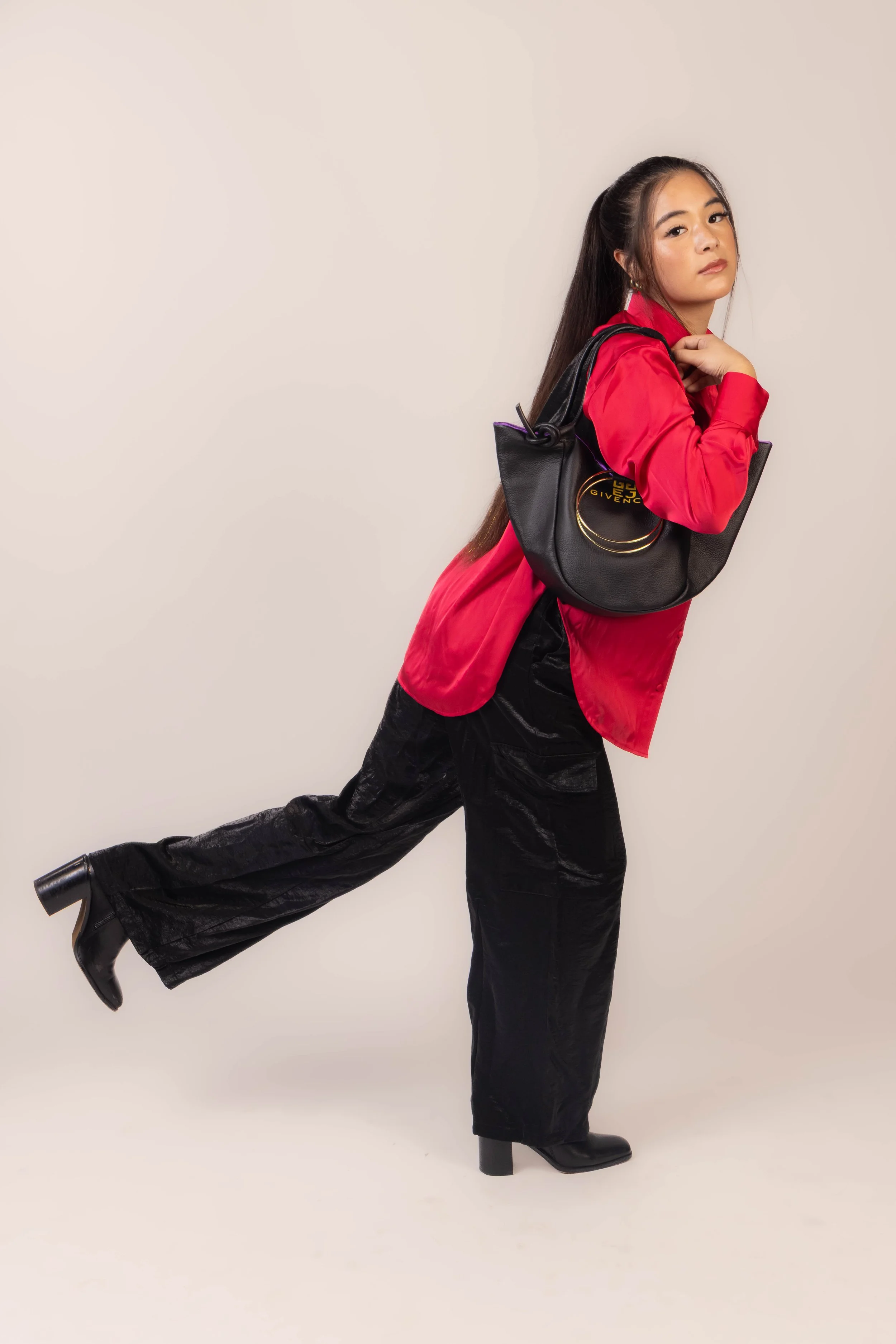 A woman with long dark hair in a high ponytail wearing a red jacket, black pants, and black heeled boots. She is holding a black Givenchy handbag over her shoulder and standing against a plain neutral-colored background. She is posing with one leg ex