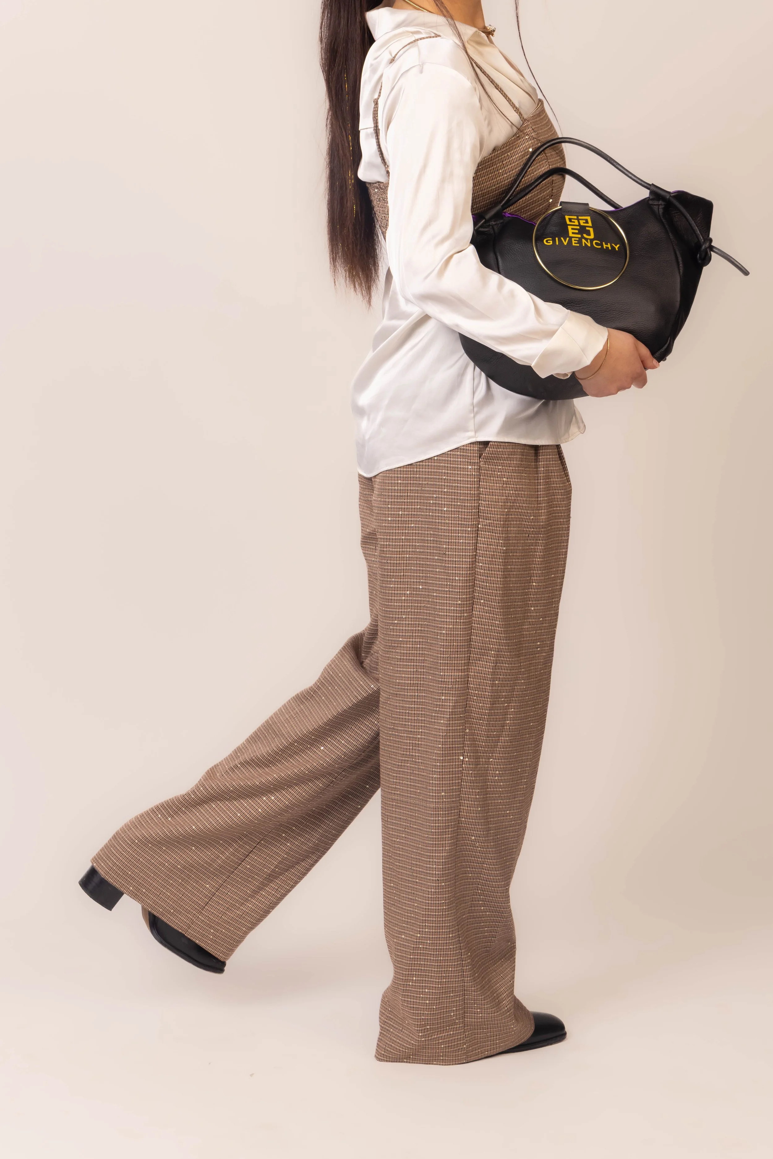 A person holding a black Givenchy handbag, dressed in brown plaid wide-leg pants, a white blouse, and a beige vest with long dark hair, standing against a plain light background.