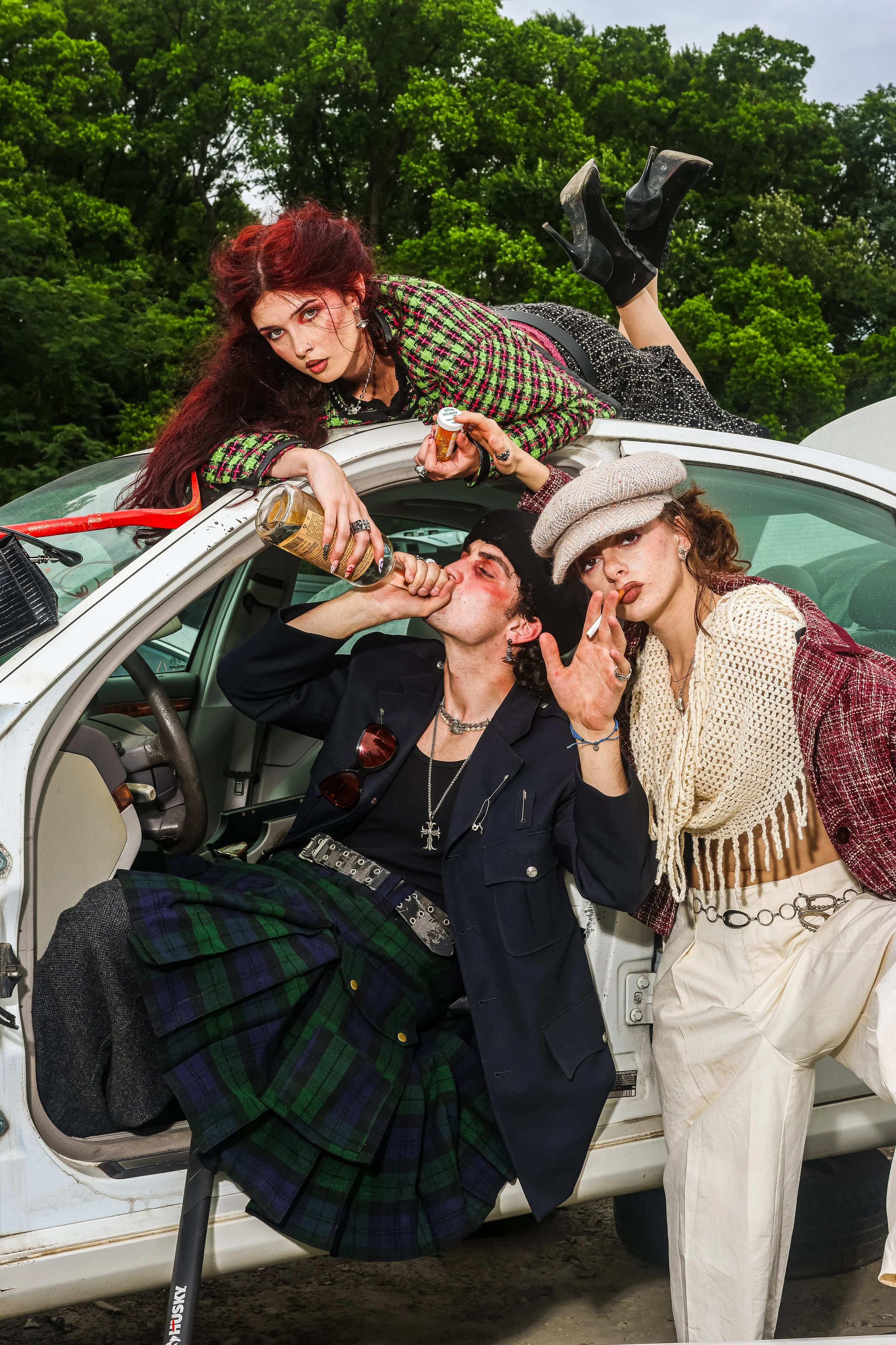 A group of young women and a man hanging out around a white car outdoors. One woman lies on top of the car, another ladles drinks, and others pose with cigarettes. They are dressed in stylish, colorful clothing and accessories with a backdrop of gree