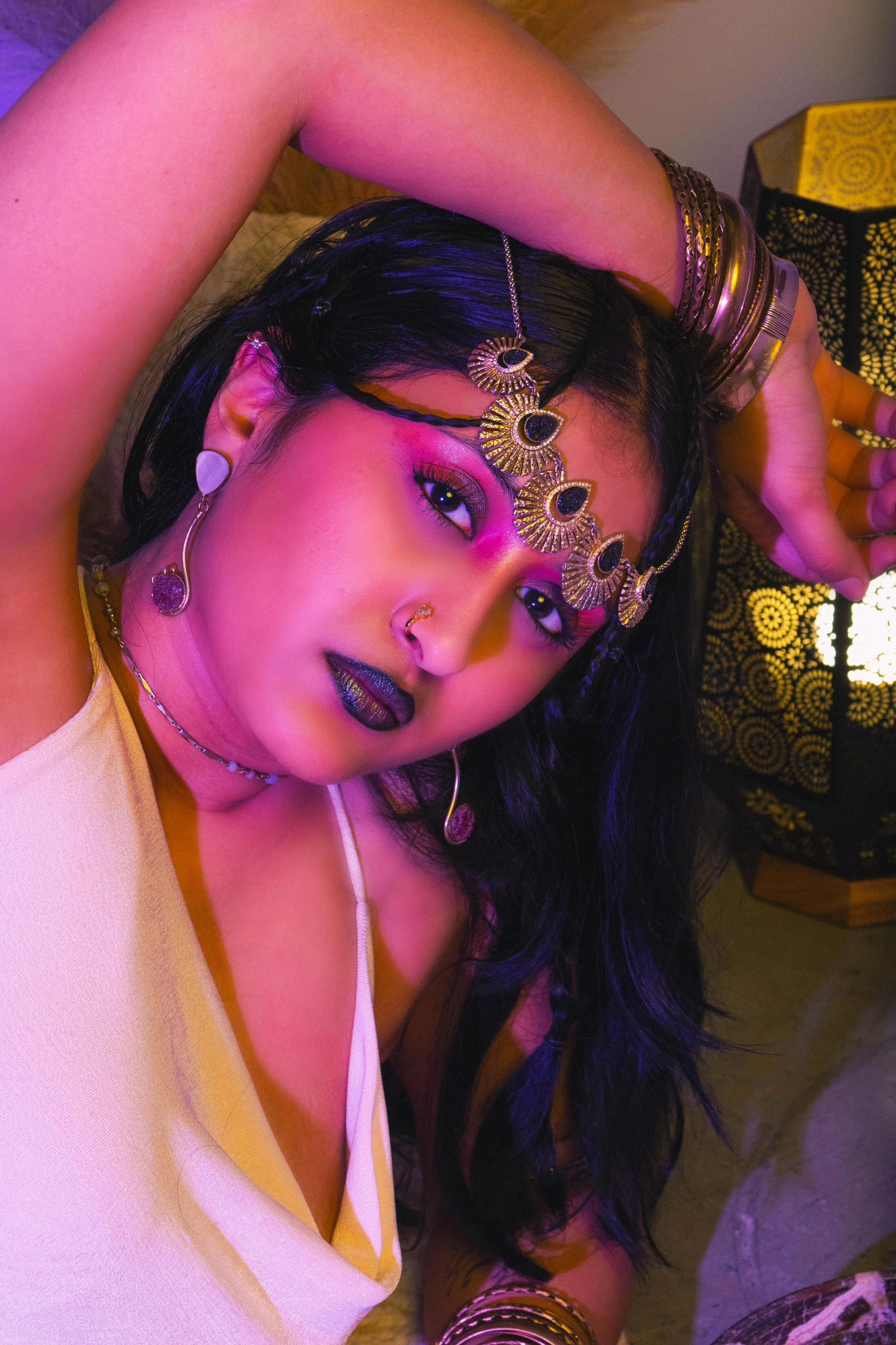 A woman with dark hair, wearing colorful makeup, jewelry, and clothing in a setting with decorative lighting.