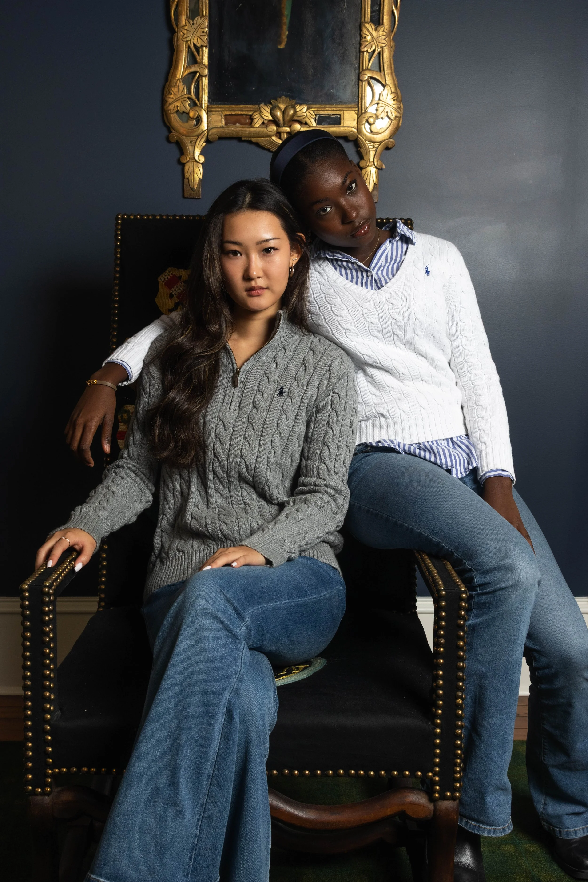 Two young women sitting together on a black and gold ornate armchair; one woman with long wavy dark hair wearing a beige cable knit sweater and blue jeans, the other woman with dark skin, short hair, wearing a white cable knit sweater with a blue and