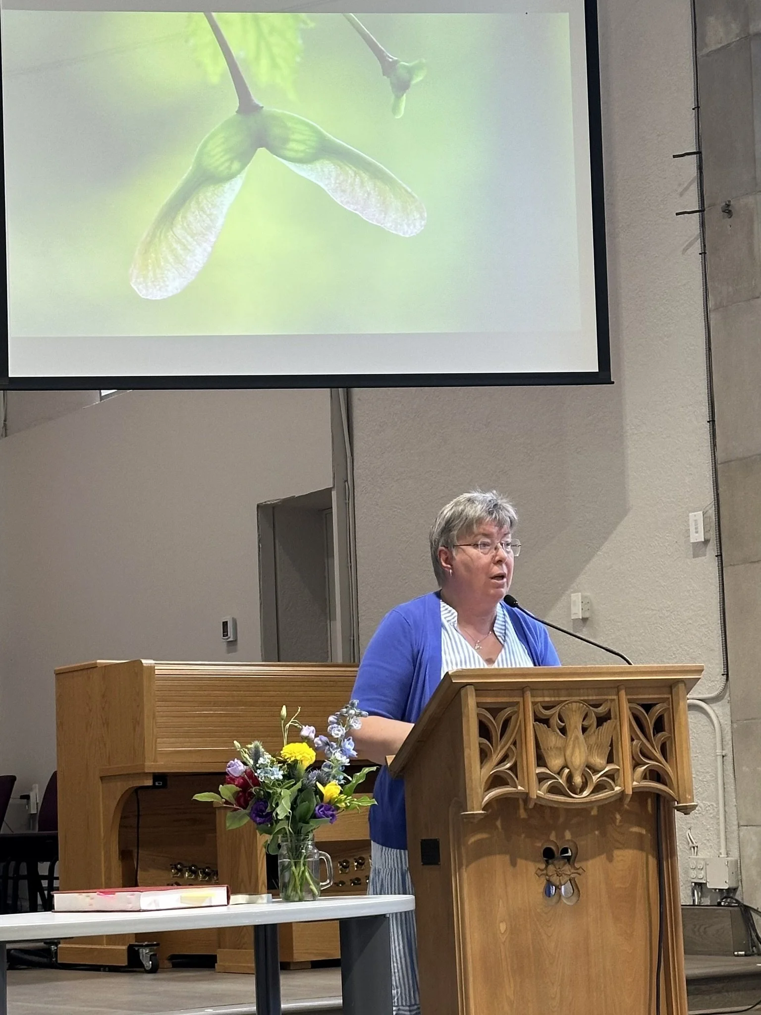 Rev. Dr. Jones speaking at the lectern, presenting her book "Midrashic Imagination." A slide projection shows sycamore seeds.
