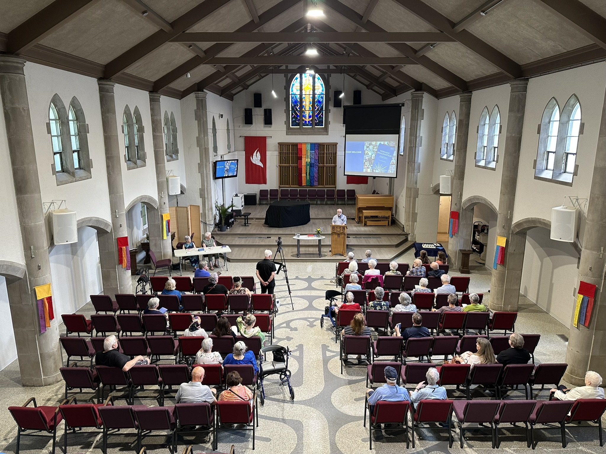 Balcony view of the presentation by Rev. Dr. Elisabeth Jones at the book launch in June 2026. The sanctuary window reflects the bright colours of the pentecost banners on the walls.