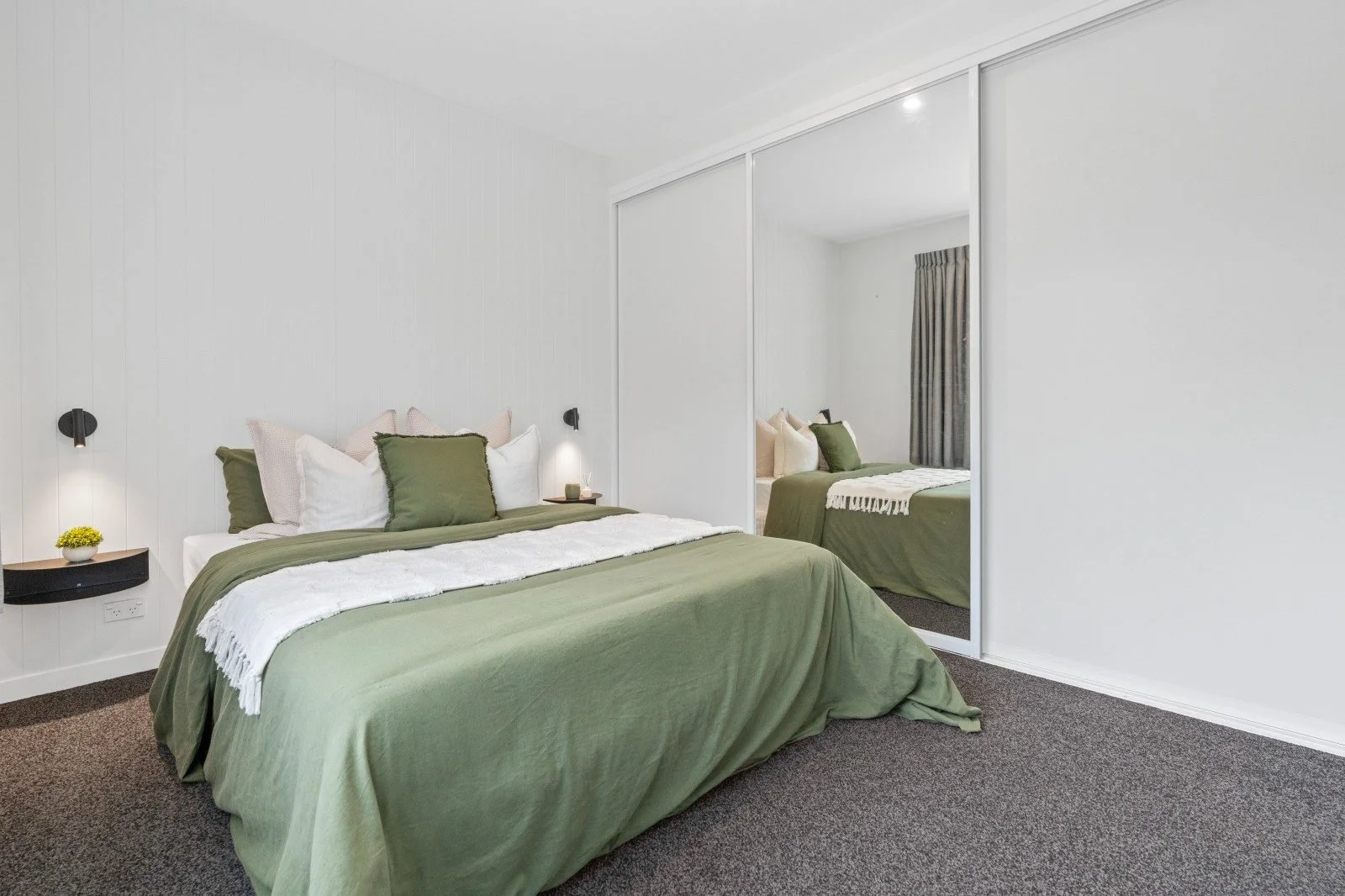 A bedroom with a bed covered in green bedding and multiple pillows, a mirrored closet door reflecting the bed, wall-mounted lights, a small bedside table with a candle and decoration, and a window with dark curtains.