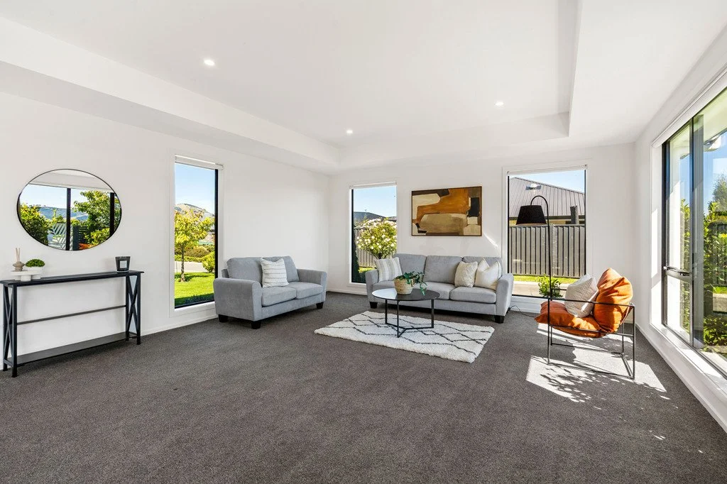 Dunedin Property Staging - Bright living room with white walls, large windows showing greenery outside, grey sofa, orange and beige armchair, modern black coffee table, mirror, abstract artwork, and a black console table.