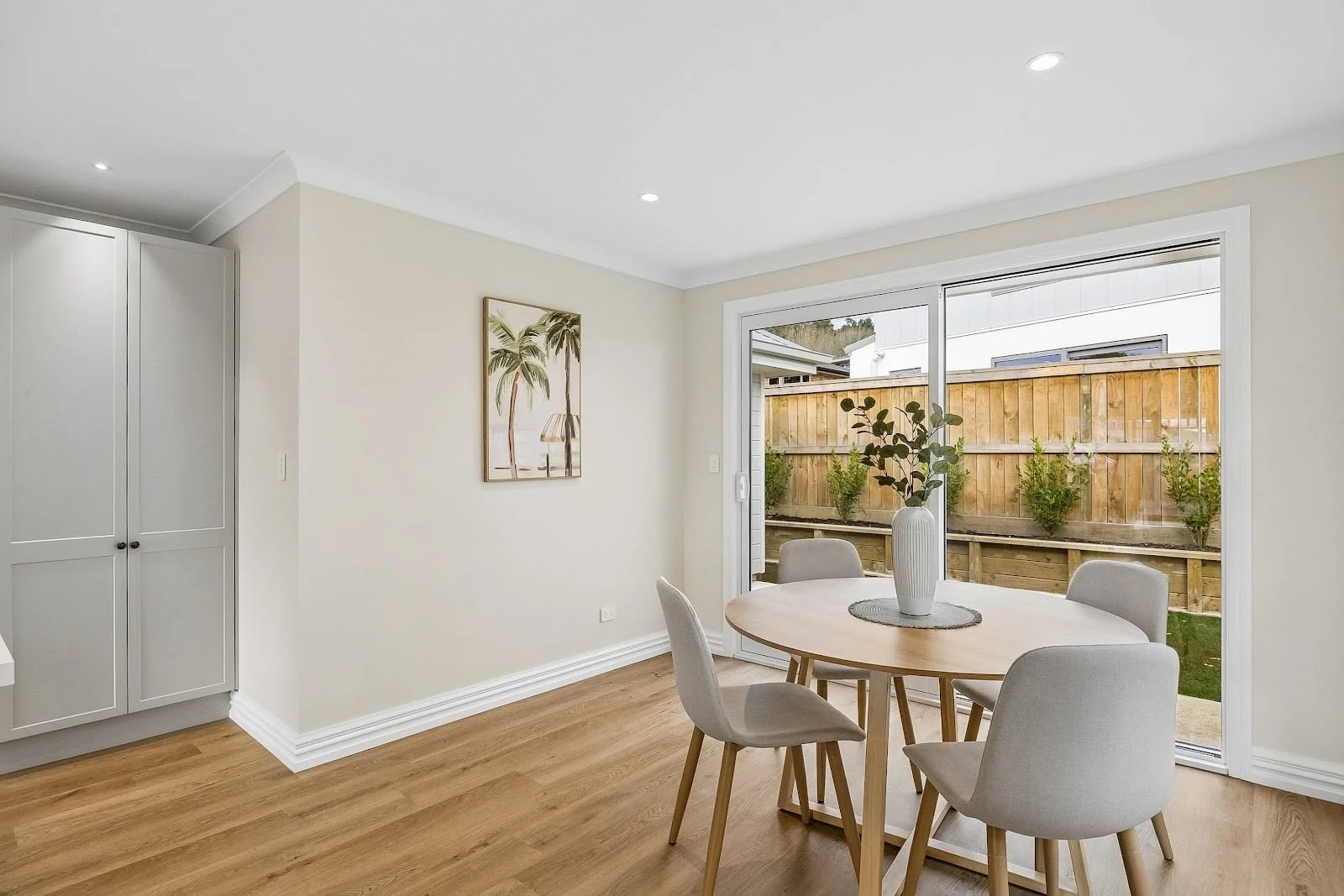 Interior of a dining area with a wooden table, four light-colored chairs, a white vase with greenery, and large sliding glass doors leading to an outdoor wooden fenced yard with small plants.