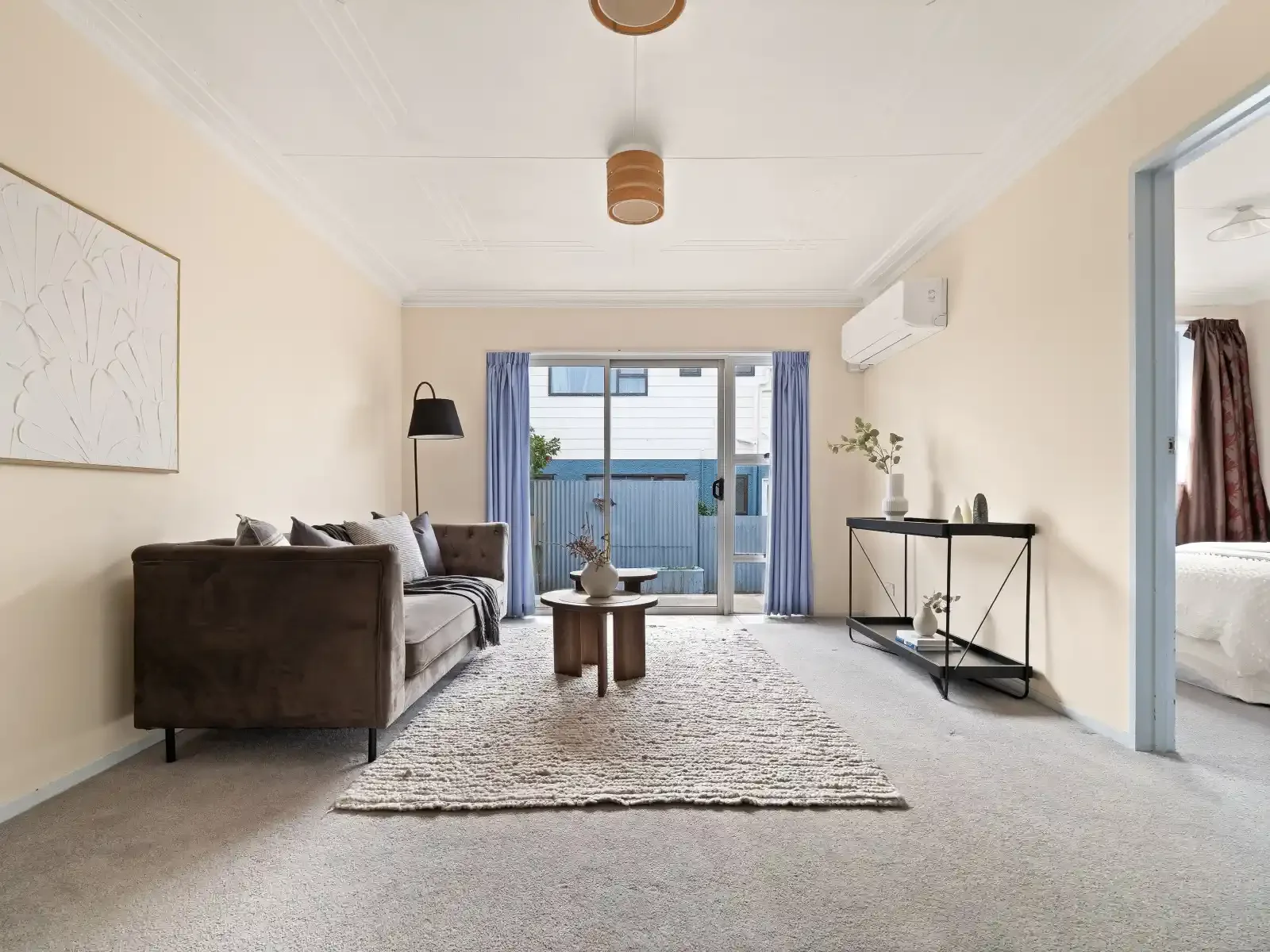 Living room with beige walls, a brown sofa with pillows, a black floor lamp, a wooden coffee table with a decorative vase, a rug, a black console table with vases, and a sliding glass door with blue curtains leading outside. Part of a bedroom with wh