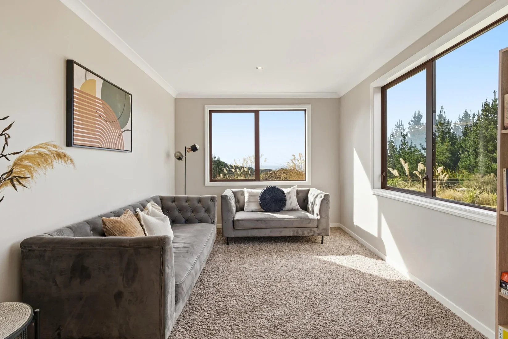 Cozy living room with two gray sofas, large windows showing outdoor greenery, modern art on the wall, and a beige carpet.