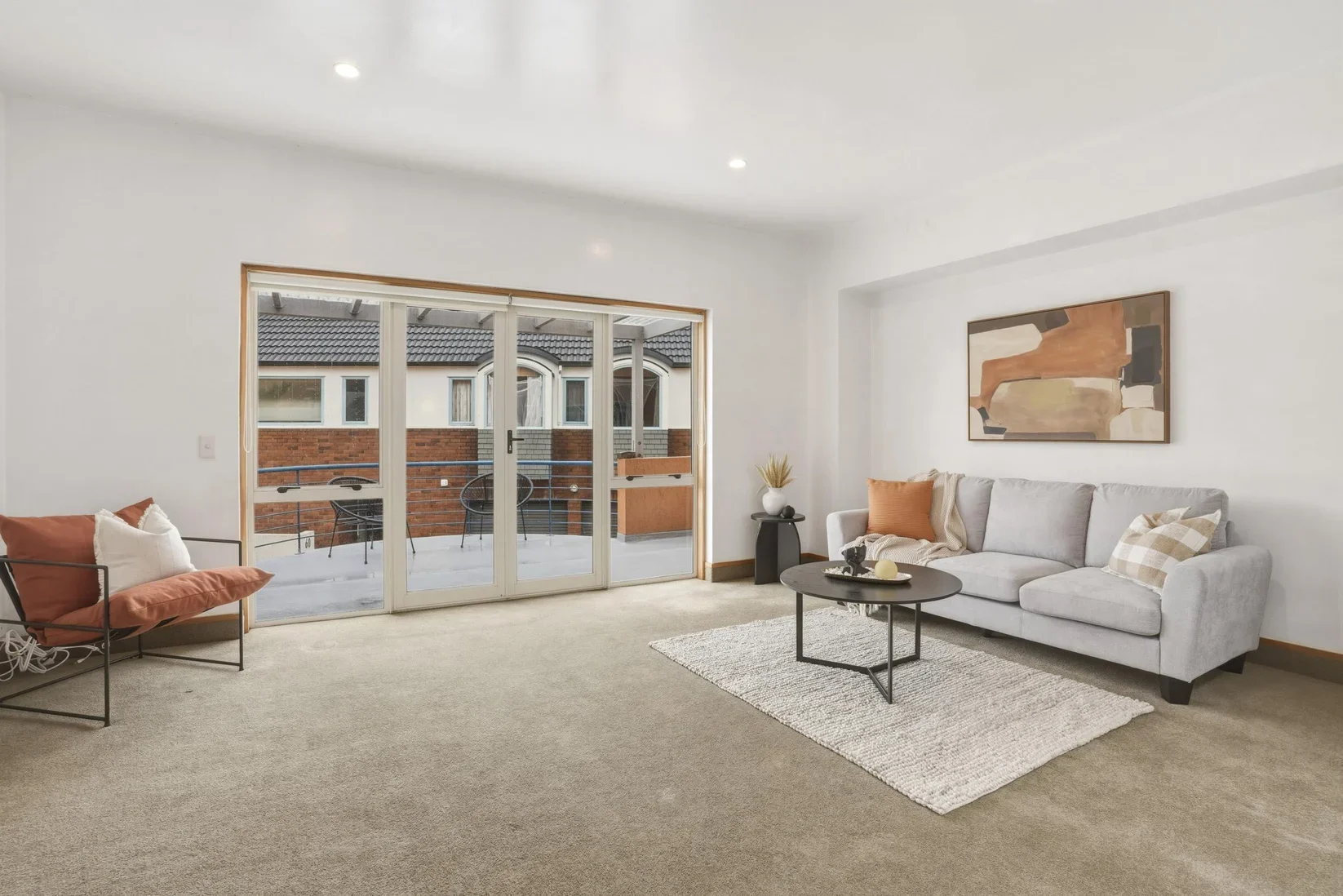 Dunedin Property Staging - Living room with a grey couch, orange and plaid pillows, a black coffee table, a side table with a vase, an abstract artwork on the wall, a large sliding glass door leading to a balcony with outdoor chairs, a beige carpet, 