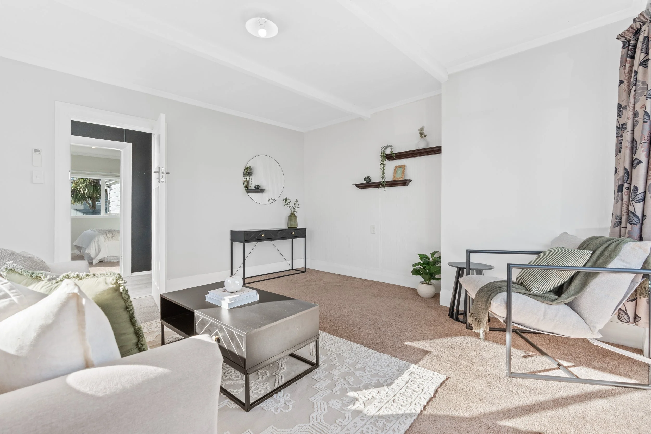 Living room with white walls, beige carpet, modern black and white coffee table, white couch with green pillows, gray armchair, and a small black side table. Decor includes floating shelves, potted plants, framed photo, round mirror, and curtains wit